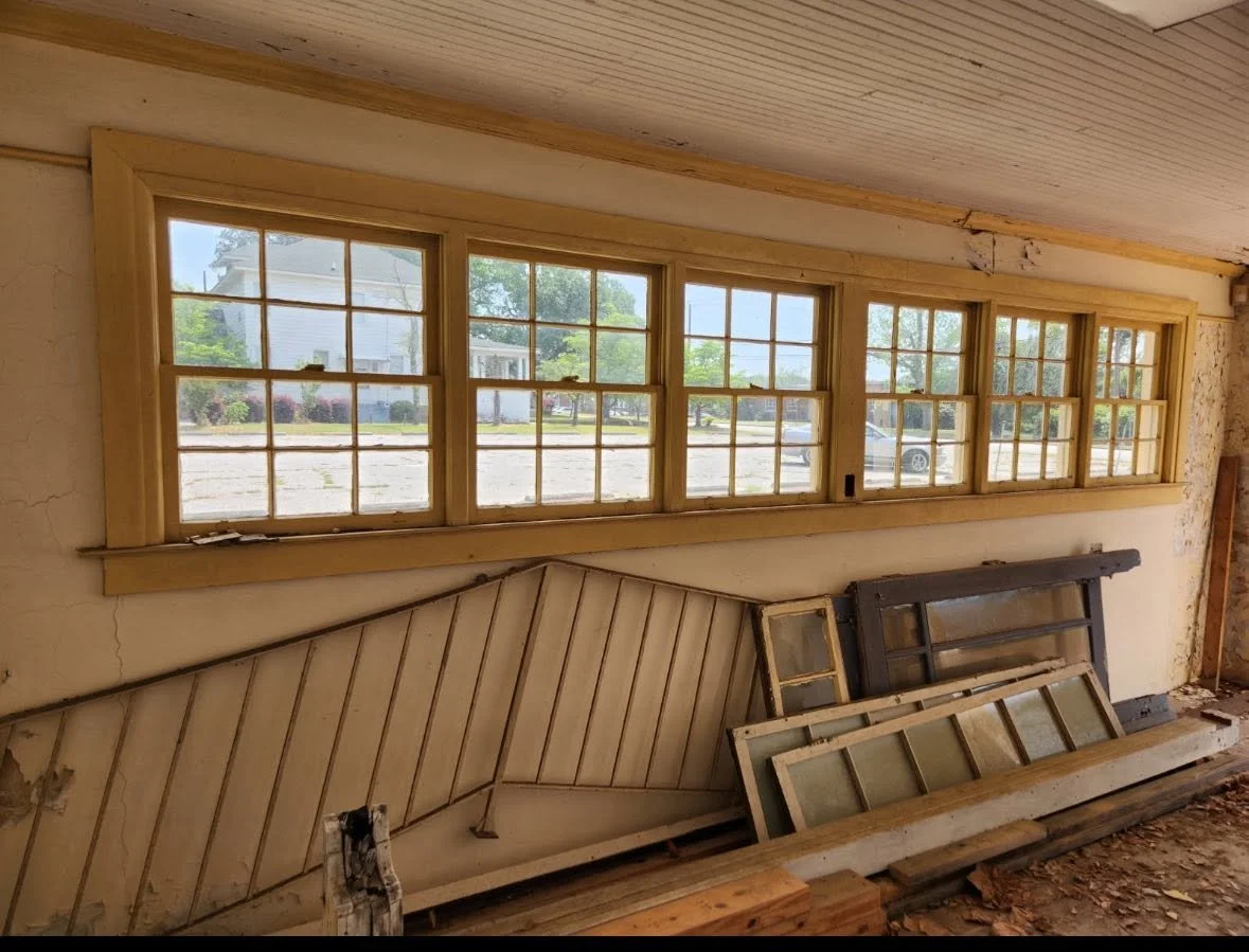Inside a room with a set of six large picture windows on the wall, and demolition debris including parts of window frames, wood, and dirt on the floor.