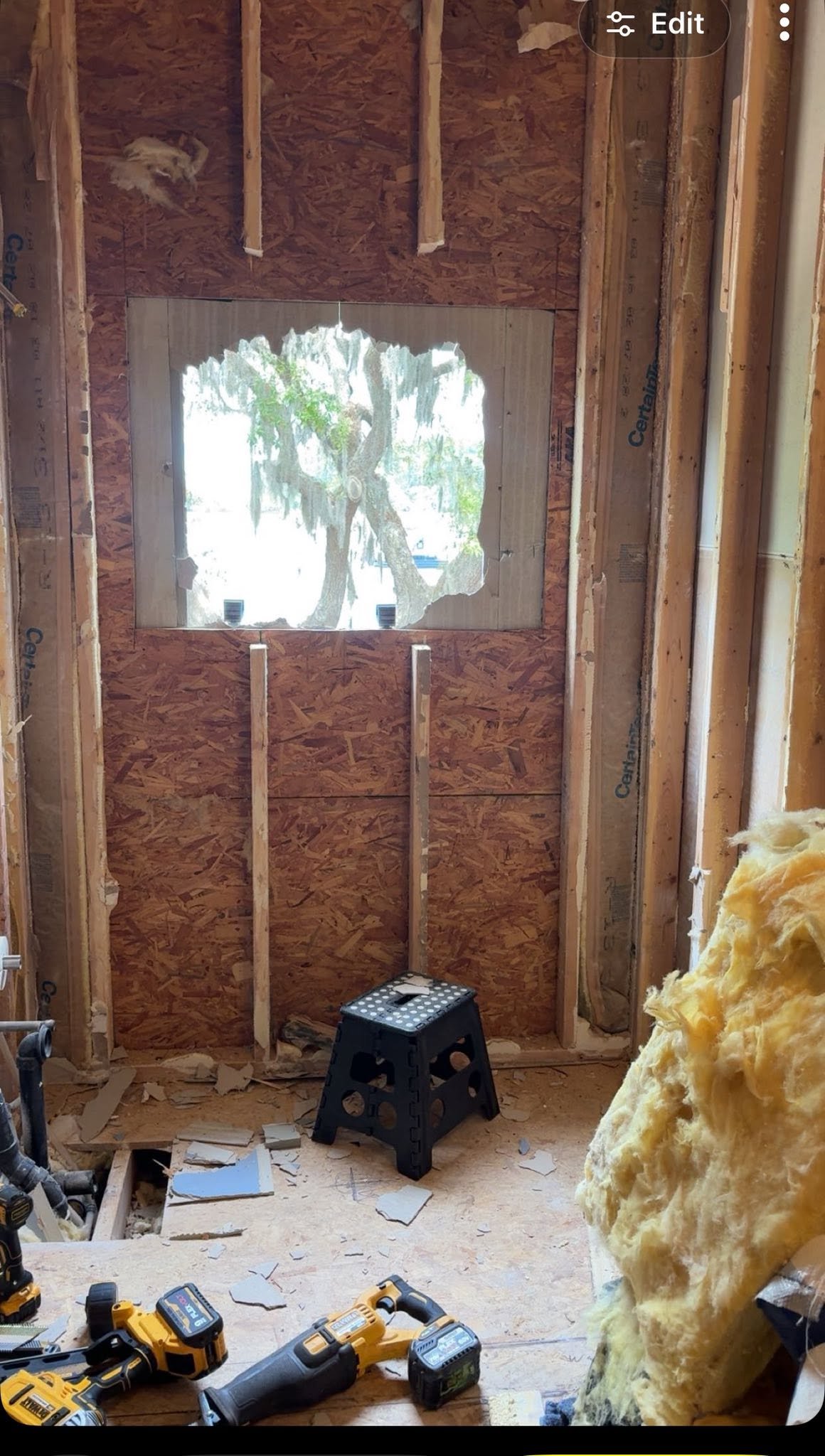 Room under construction with exposed wooden studs and insulation, a small window showing tree outside, construction tools on the floor, a plastic step stool, and torn drywall pieces.