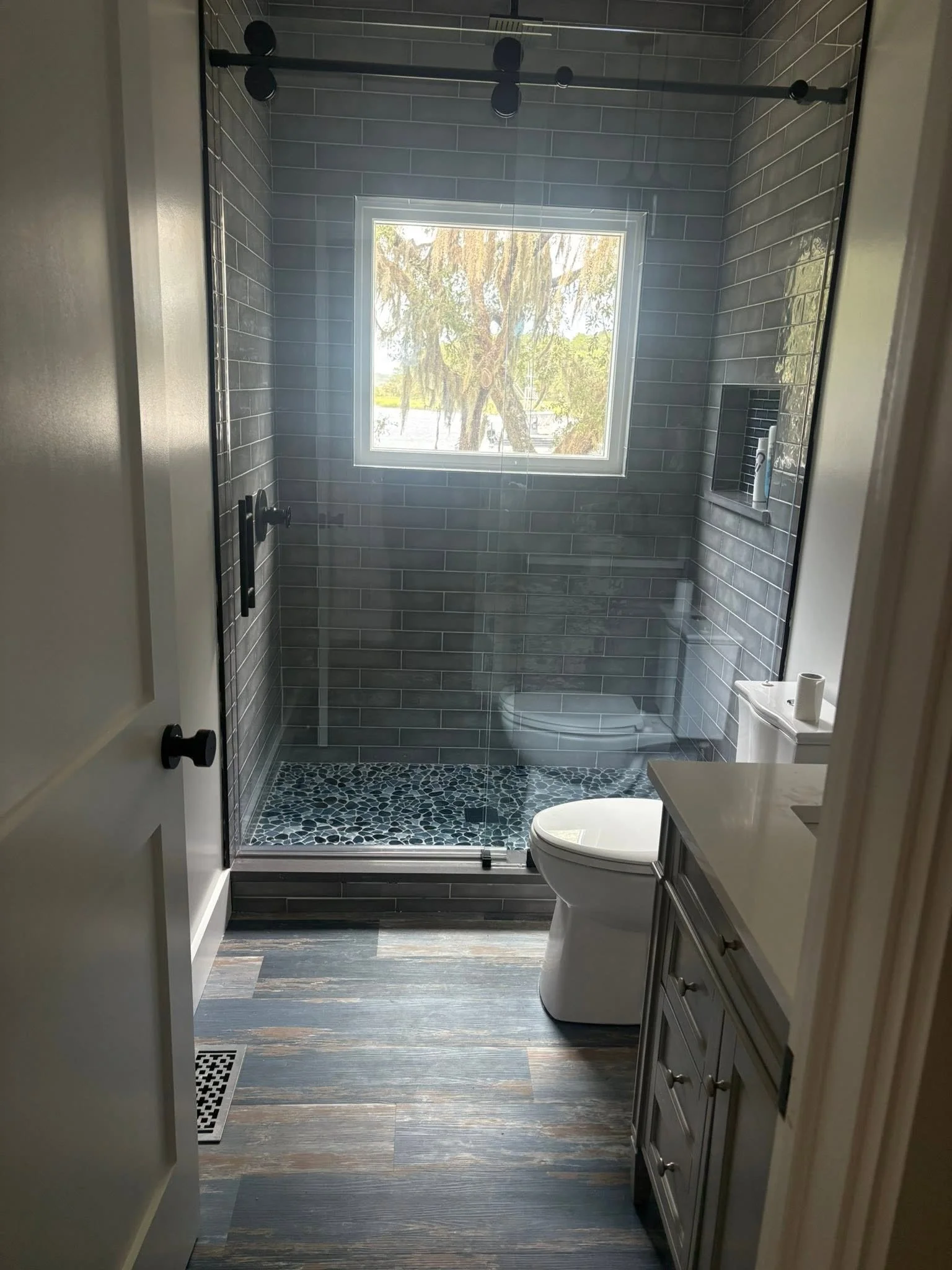 A modern bathroom with a glass shower enclosure, gray tiled walls, and a window showing trees outside. There is a tile niche with toiletries and a toilet in the foreground. The bathroom has wood-like flooring and a vanity with a countertop.