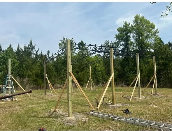 Construction scaffolding with wooden supports on a grassy field, surrounded by trees, with a ladder and tools nearby.