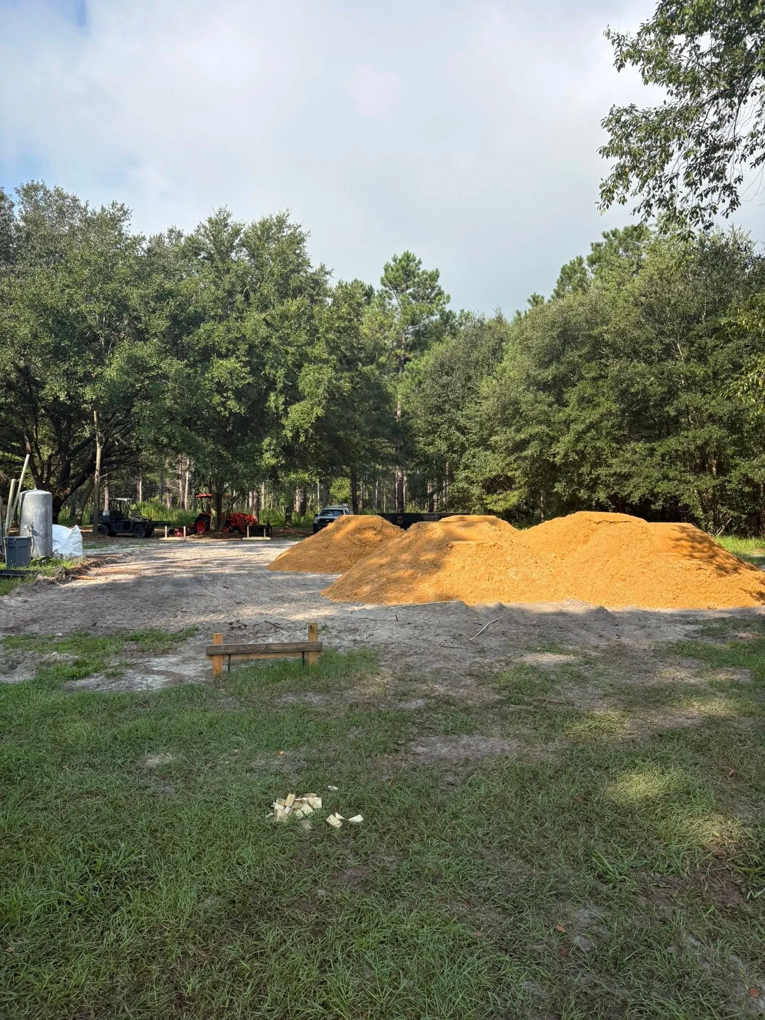A construction site with two large piles of yellow dirt, surrounded by green trees and grass.