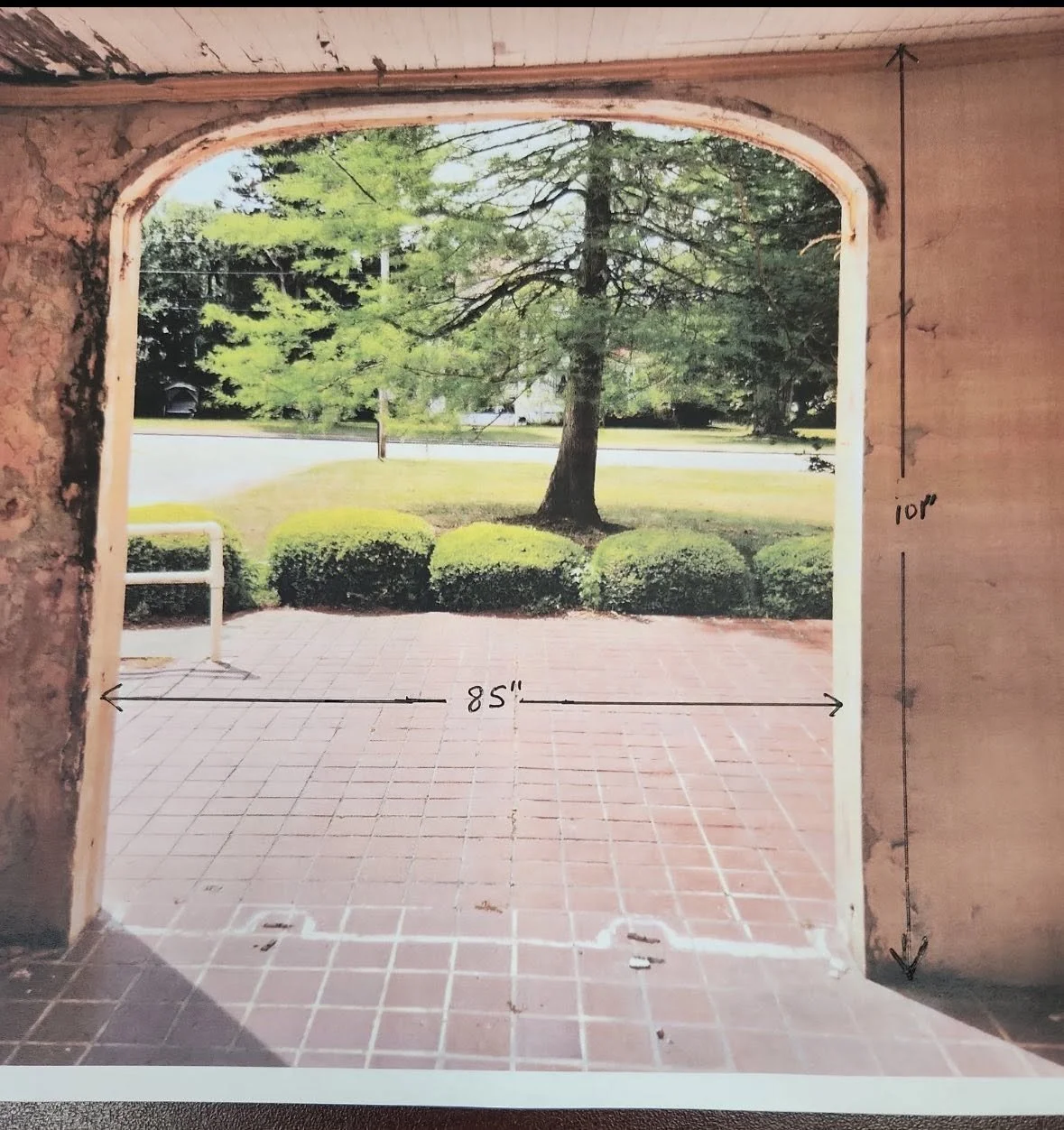 A photo taken from inside a structure looking out through a large arched opening. The image shows the dimensions of the opening — 85 inches wide and 107 inches high — handwritten on the wall on the right side. Outside, there is a brick patio directly in front, with a grassy area, bushes, and a large tree visible in the background.