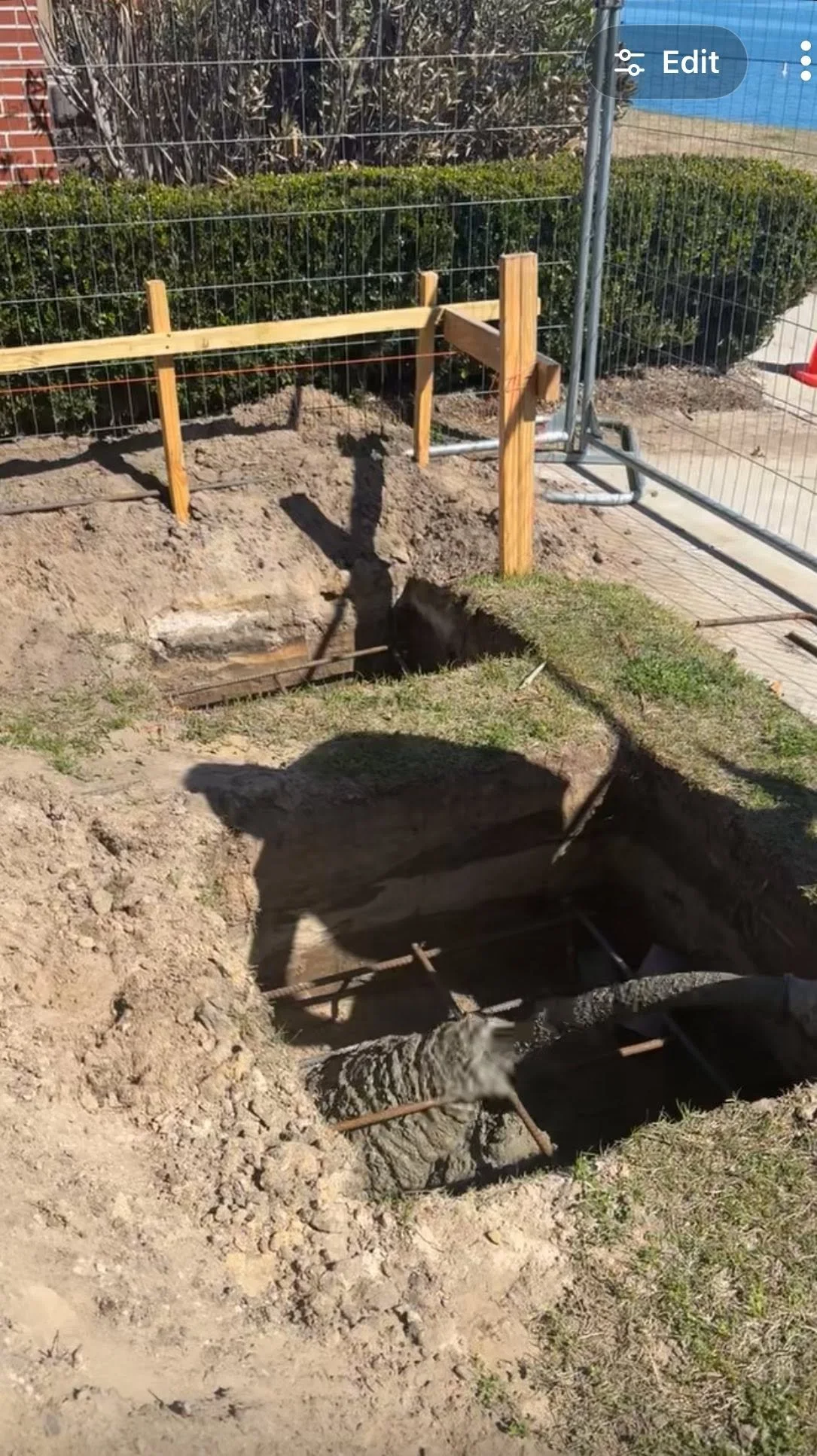 A construction site showing a deep hole in the ground with wooden framing and metal rebar, surrounded by dirt and grass.