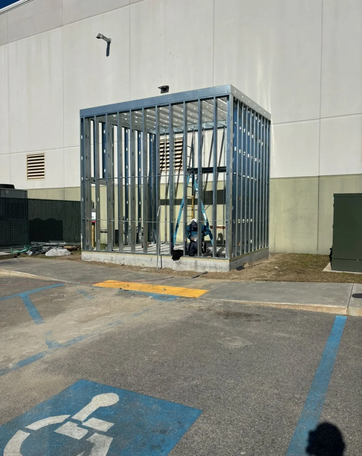 Construction worker installing the frame of a small building or enclosure outside a large building, with parking spaces in the foreground.