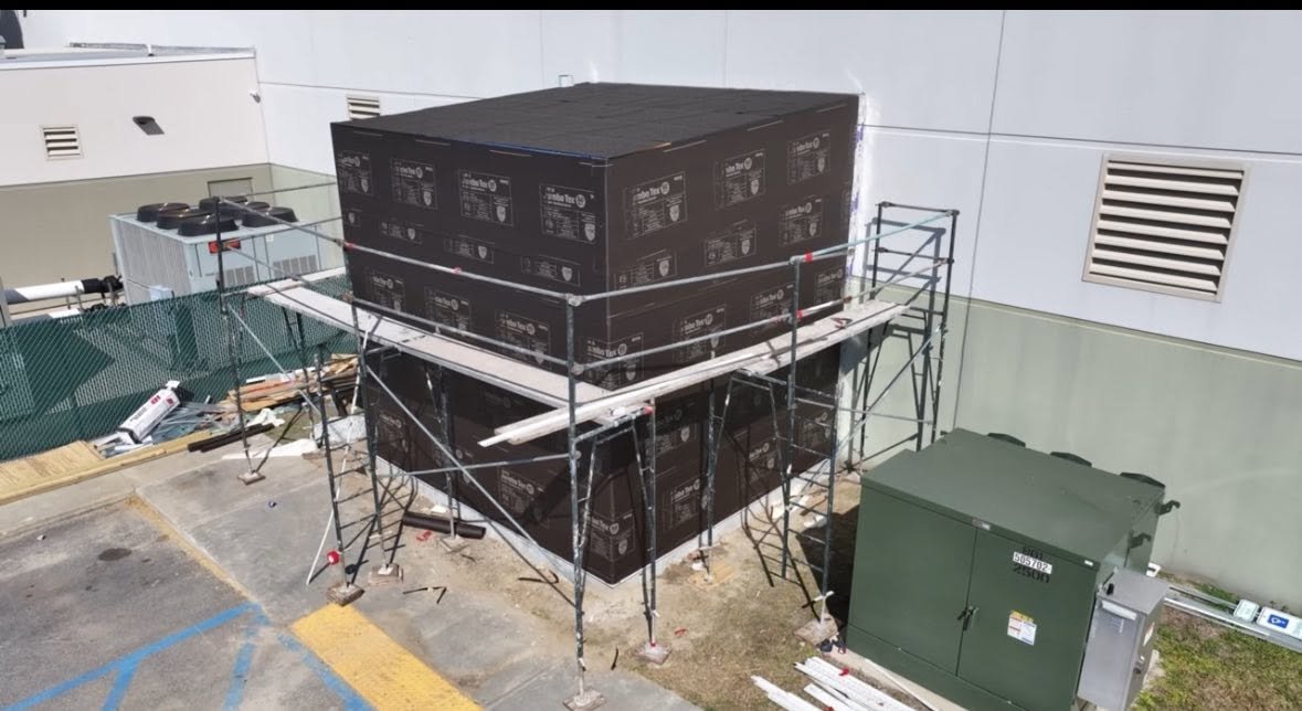 Construction scaffolding surrounds an HVAC unit outside a building, with boxes stacked above it, and construction materials on the ground nearby.