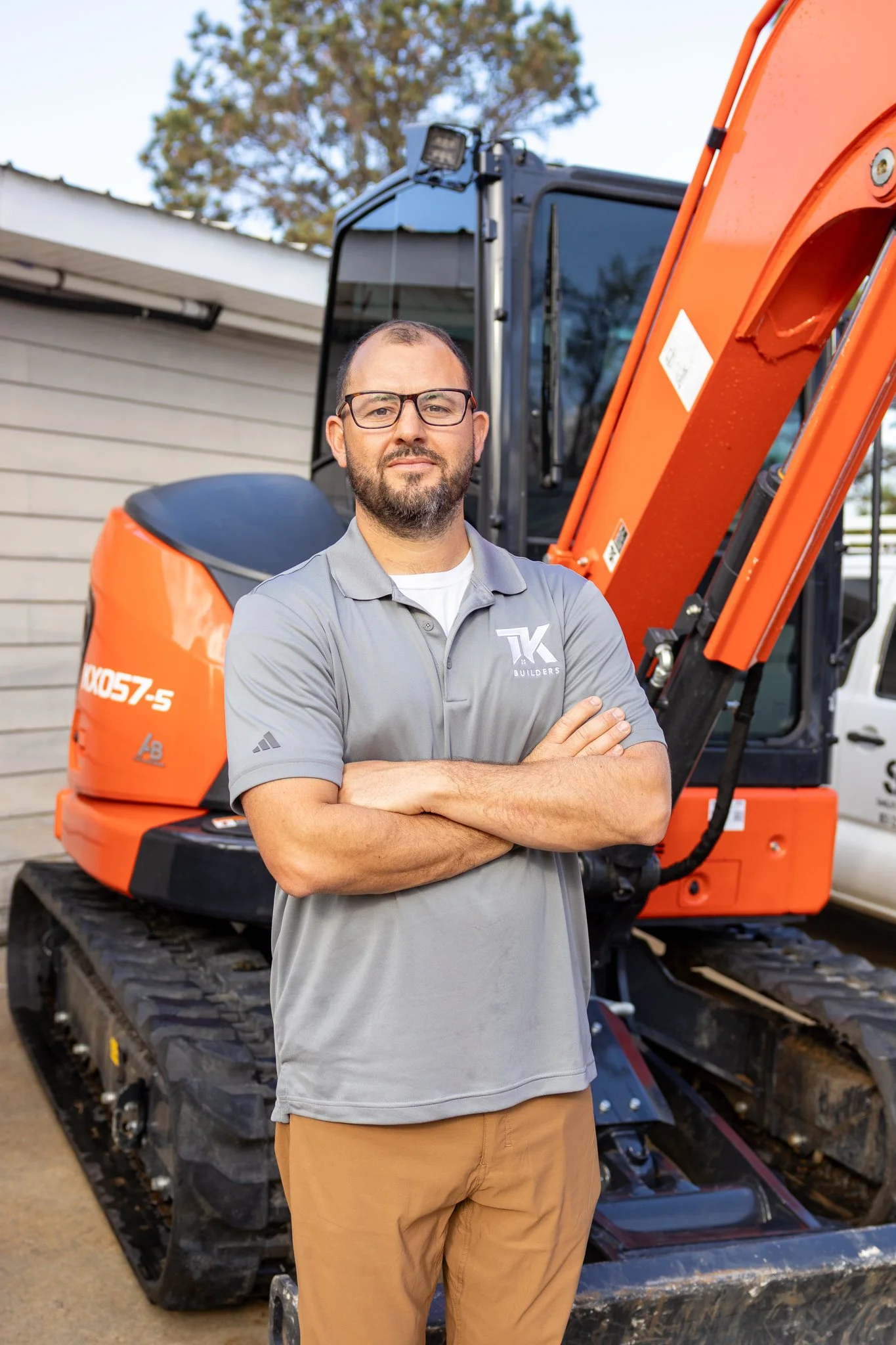 TK Builders CEO Trey Smiley wearing a gray polo and tan pants in front of an orange excavator.