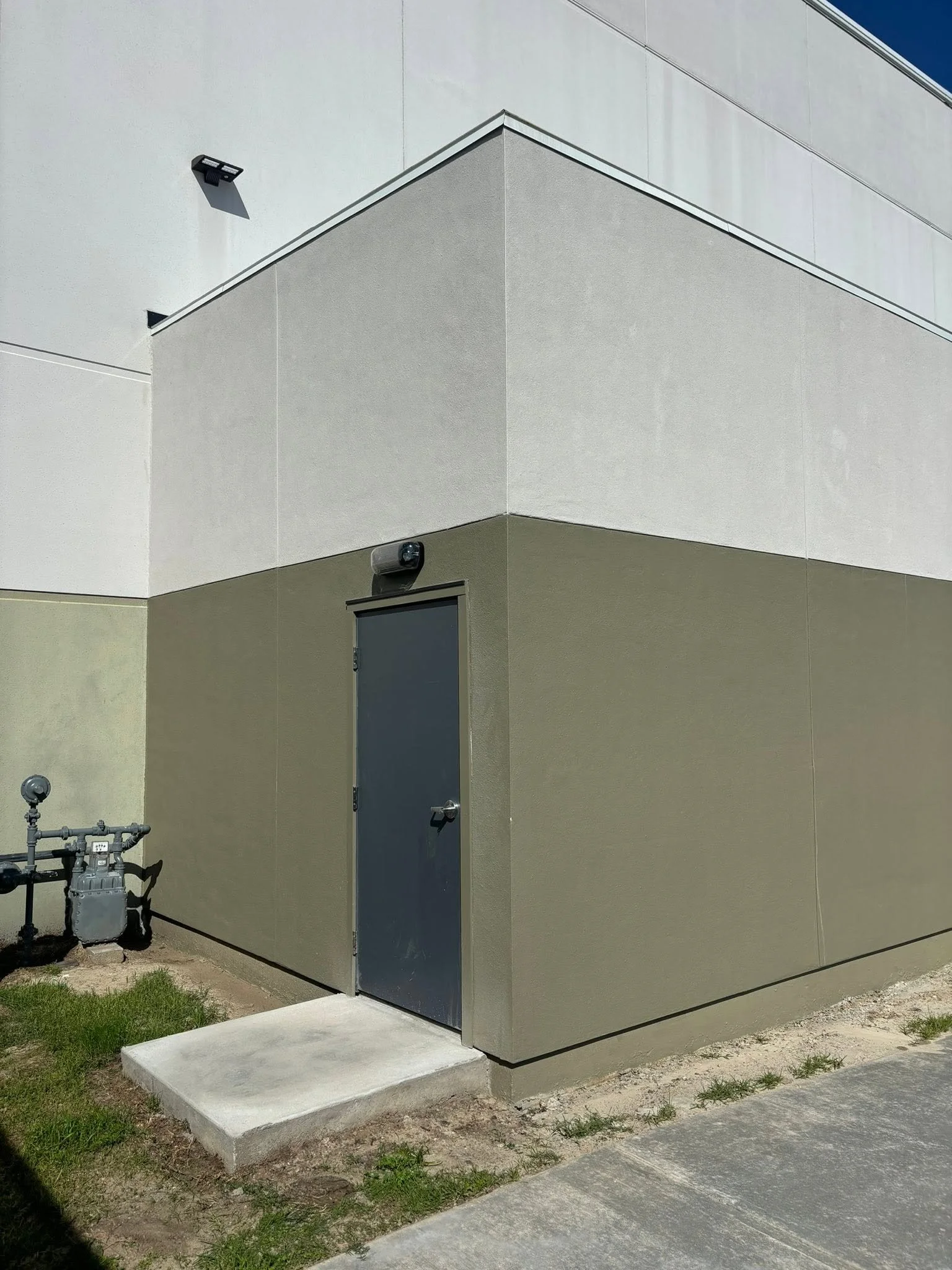 A gray utility door on a beige painted building wall, with a small white concrete step in front, next to a patch of grass and concrete pavement, with outdoor pipes and a utility meter on the left side.