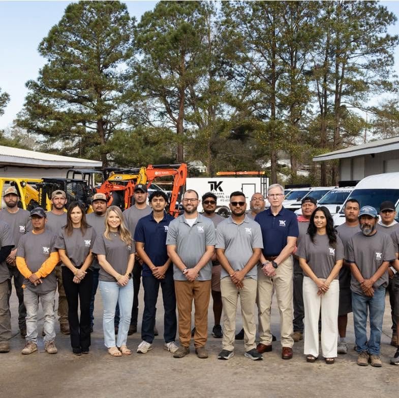 TK Builders team standing outdoors with company vehicles and construction equipment.