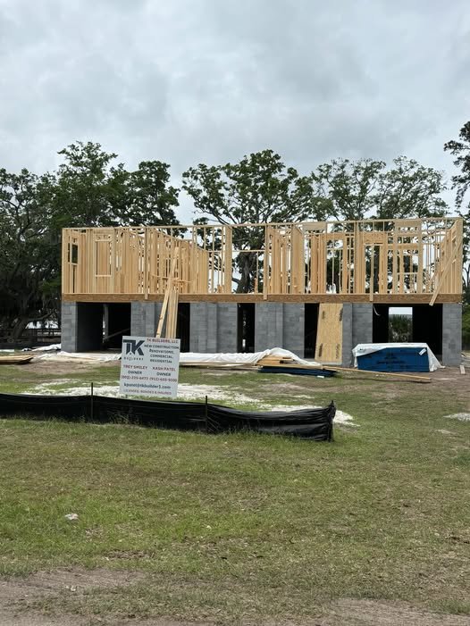 Wooden framework of a building under construction with support columns, construction sign, scattered building materials, and trees in the background.