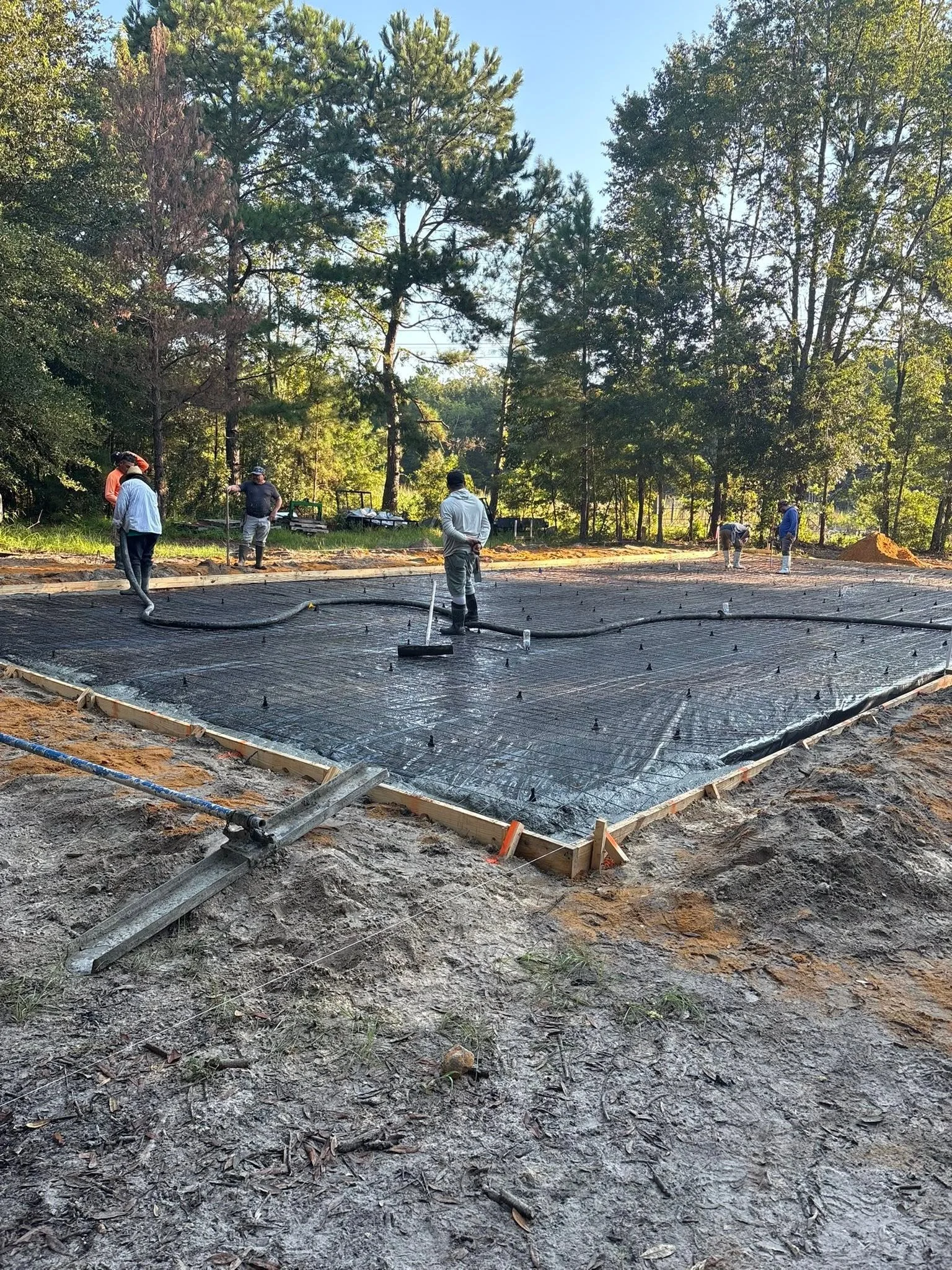 Workers laying the foundation for a building, surrounded by trees, with black plastic sheeting and wooden framing.