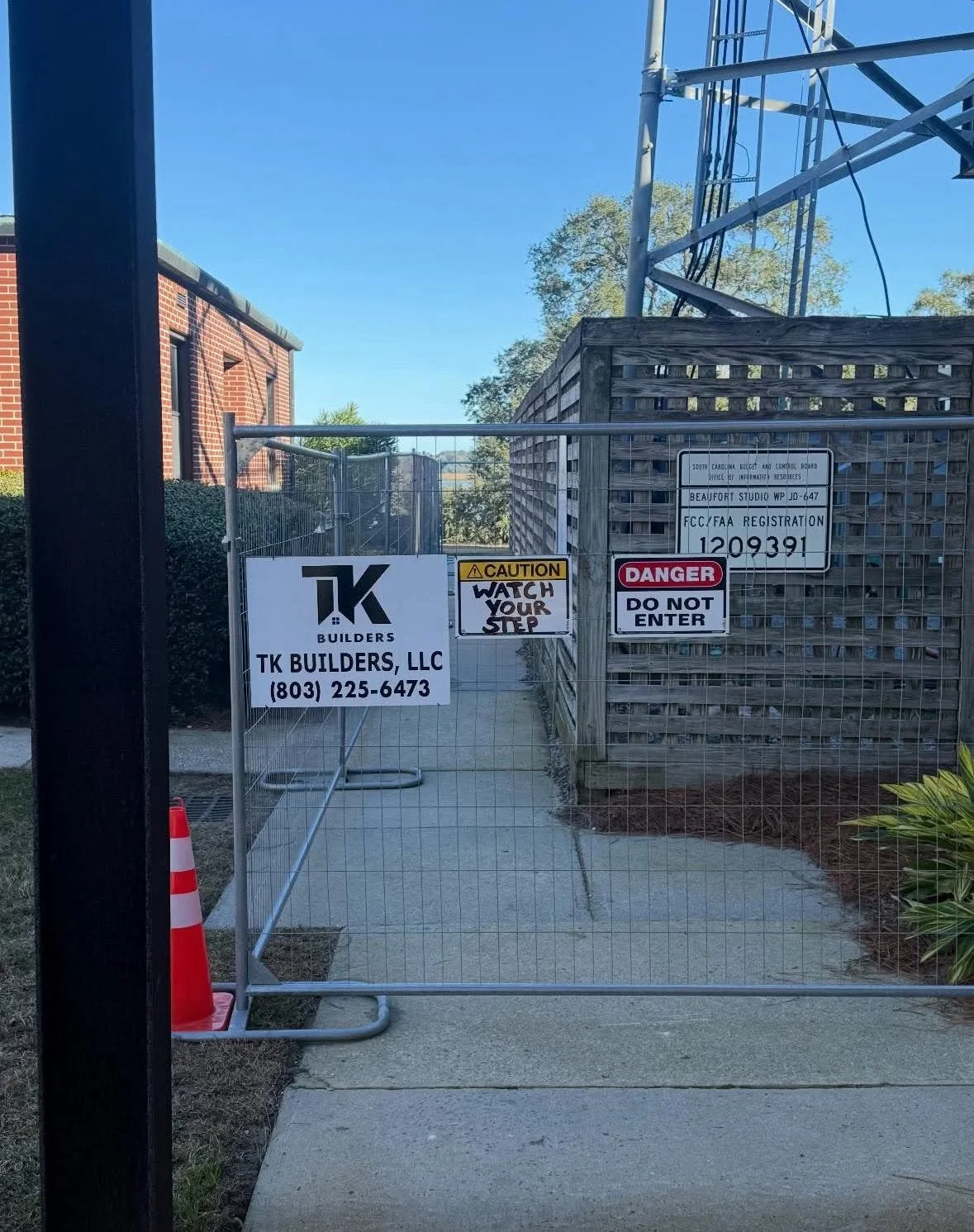 Construction site entrance with metal fence and signs, including TK Builders, a caution sign to watch your step, and a danger sign to not enter. Behind the fence is a wooden structure and a metal tower, with a sidewalk and some landscaping nearby.
