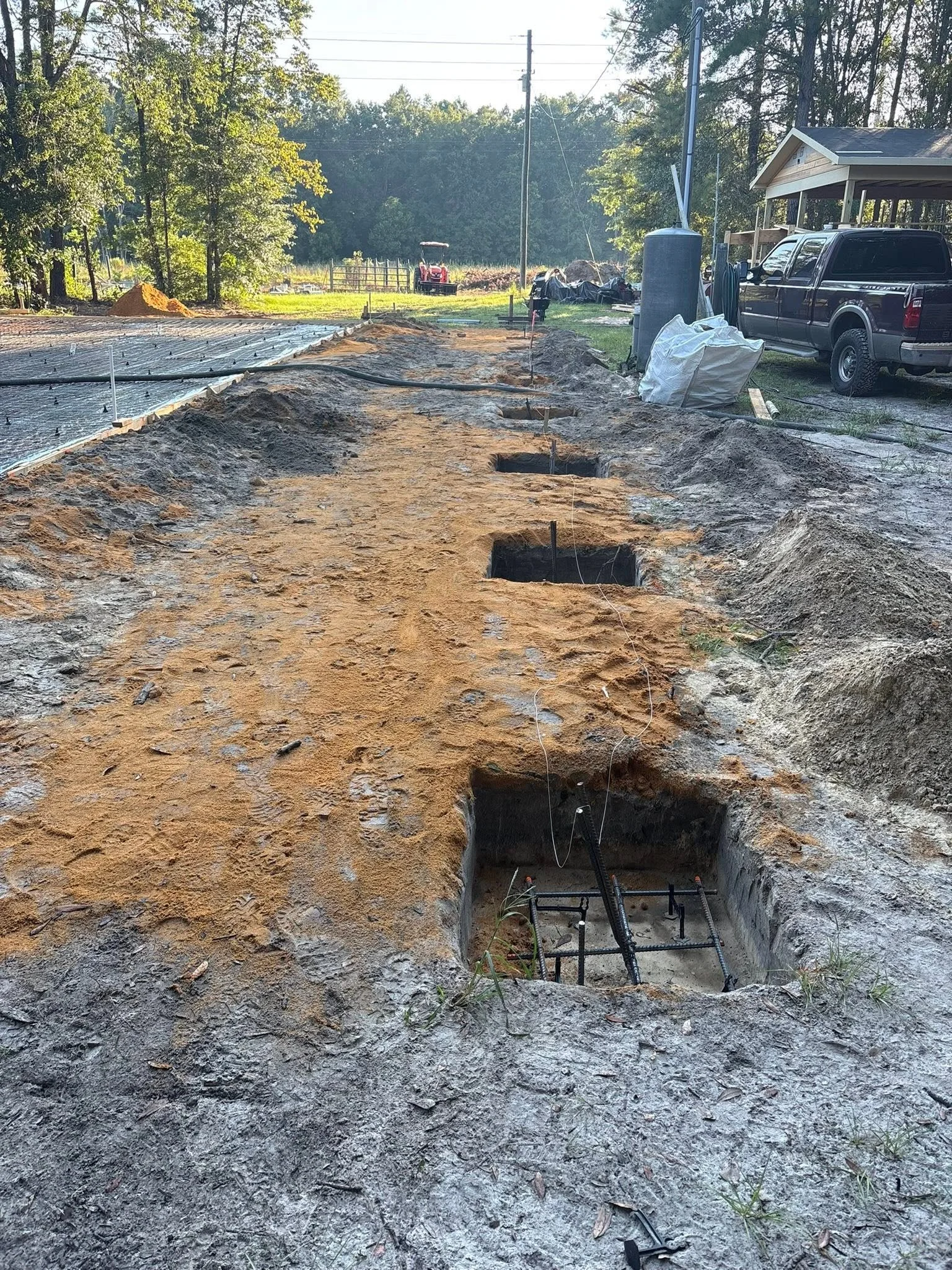 Construction site with trenches and hole dug in the ground, plumbing pipes are visible, and equipment like a pickup truck, a tractor, and construction supplies are in the background.