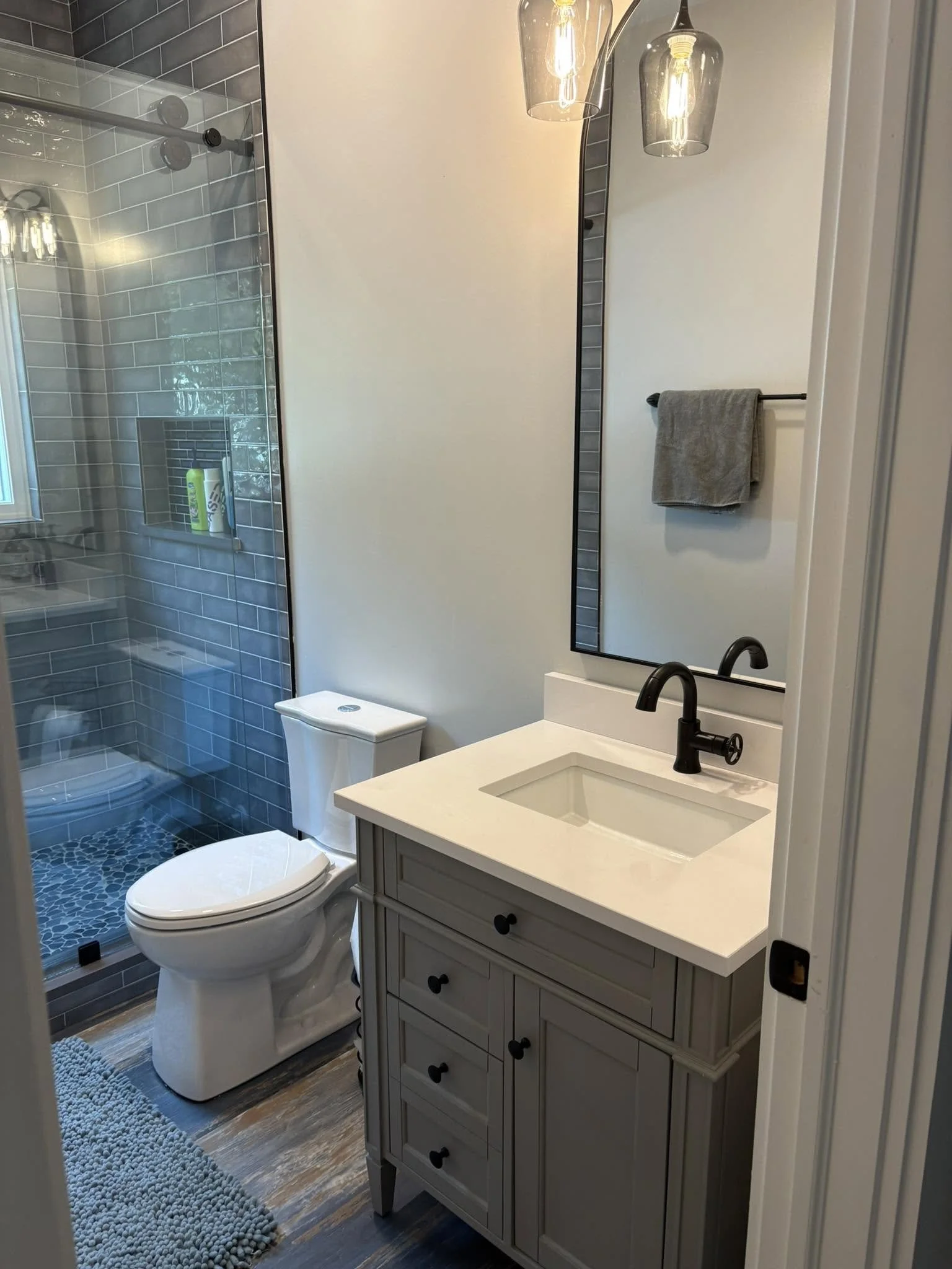 A modern bathroom with a walk-in shower with gray tile walls and a glass door, a white toilet, a gray bathroom vanity with drawers, a white countertop with an under-mount sink, a black faucet, a mirror, a beige towel on a black towel bar, a hanging light fixture with three glass shades, and a gray textured bath mat.