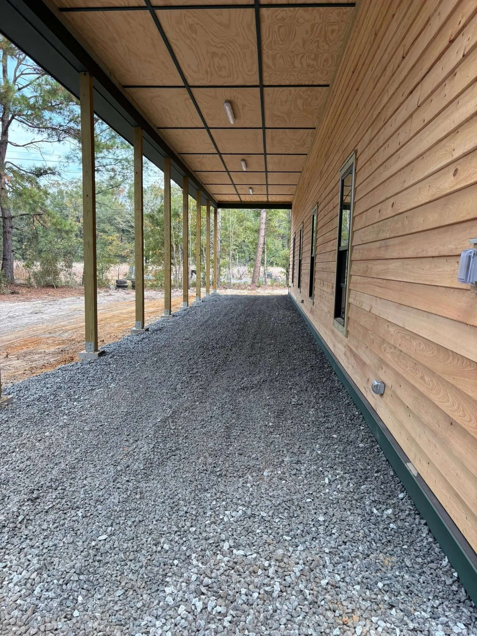Side view of a house with wooden siding, overlooking a gravel porch or walkway, with trees visible in the background, and unfinished construction on the porch ceiling.