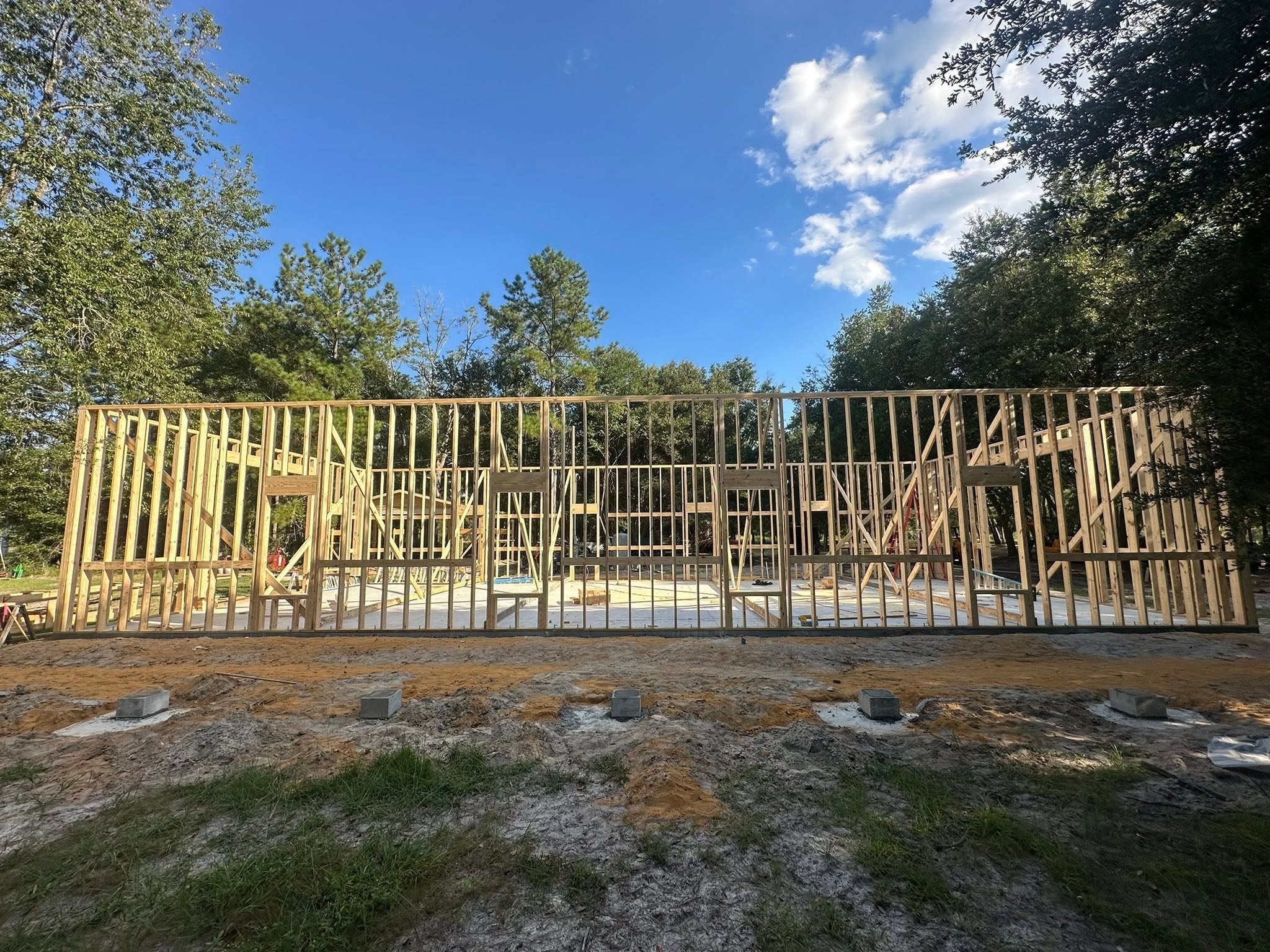 Wooden framing for a building under construction in a wooded area, with a bright blue sky and some clouds overhead.