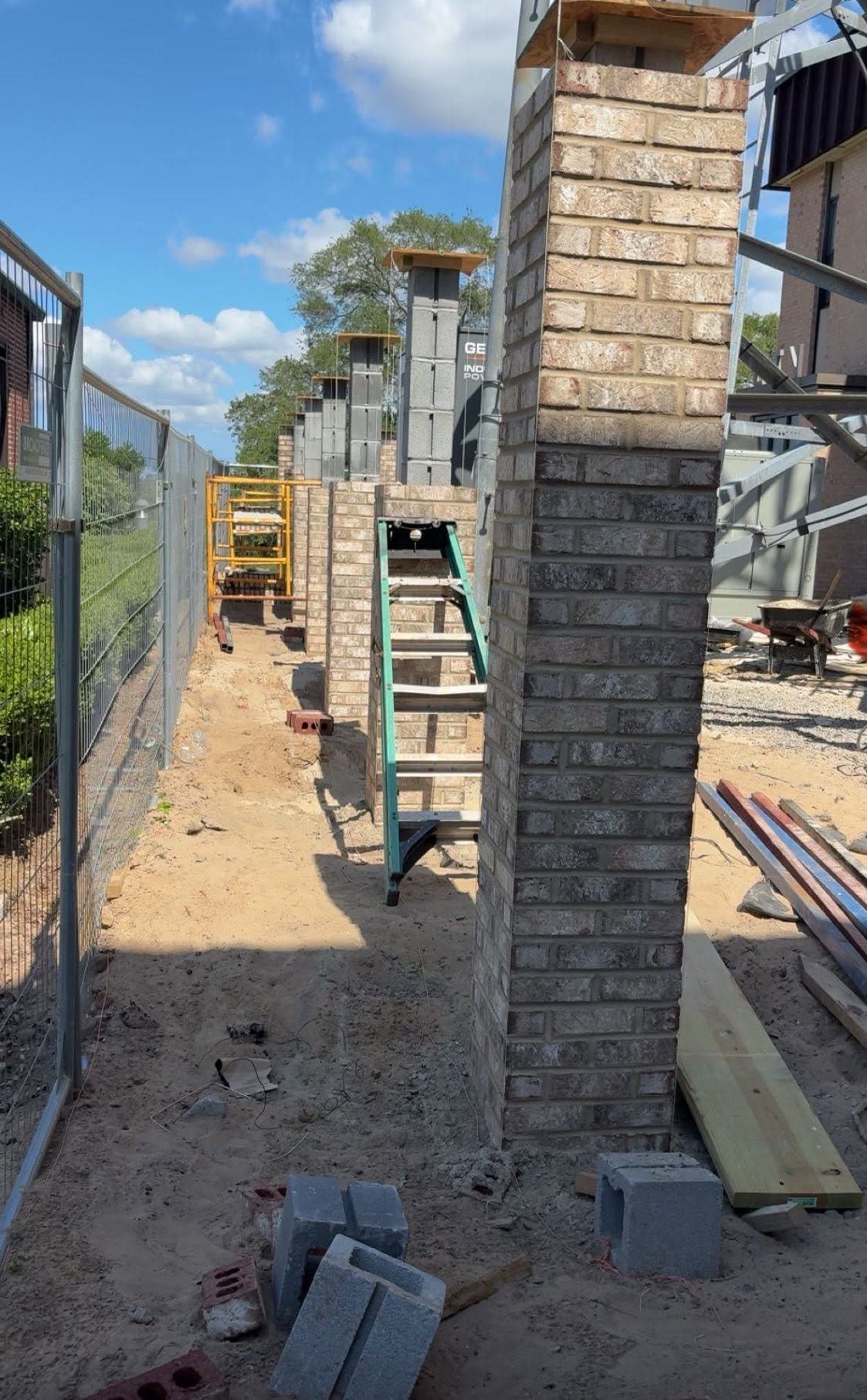 Construction site with brick pillars, a ladder, and construction materials on dirt ground, with a metal fence on the left, under a partly cloudy sky.