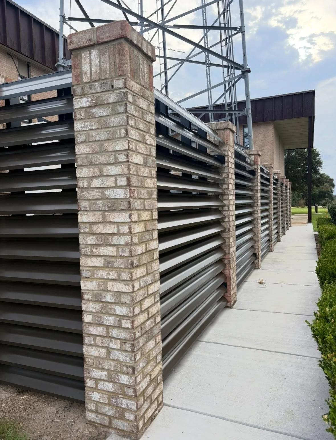 Exterior of a building with brick pillars and metal louvered vents, a sidewalk, and a landscaped lawn.
