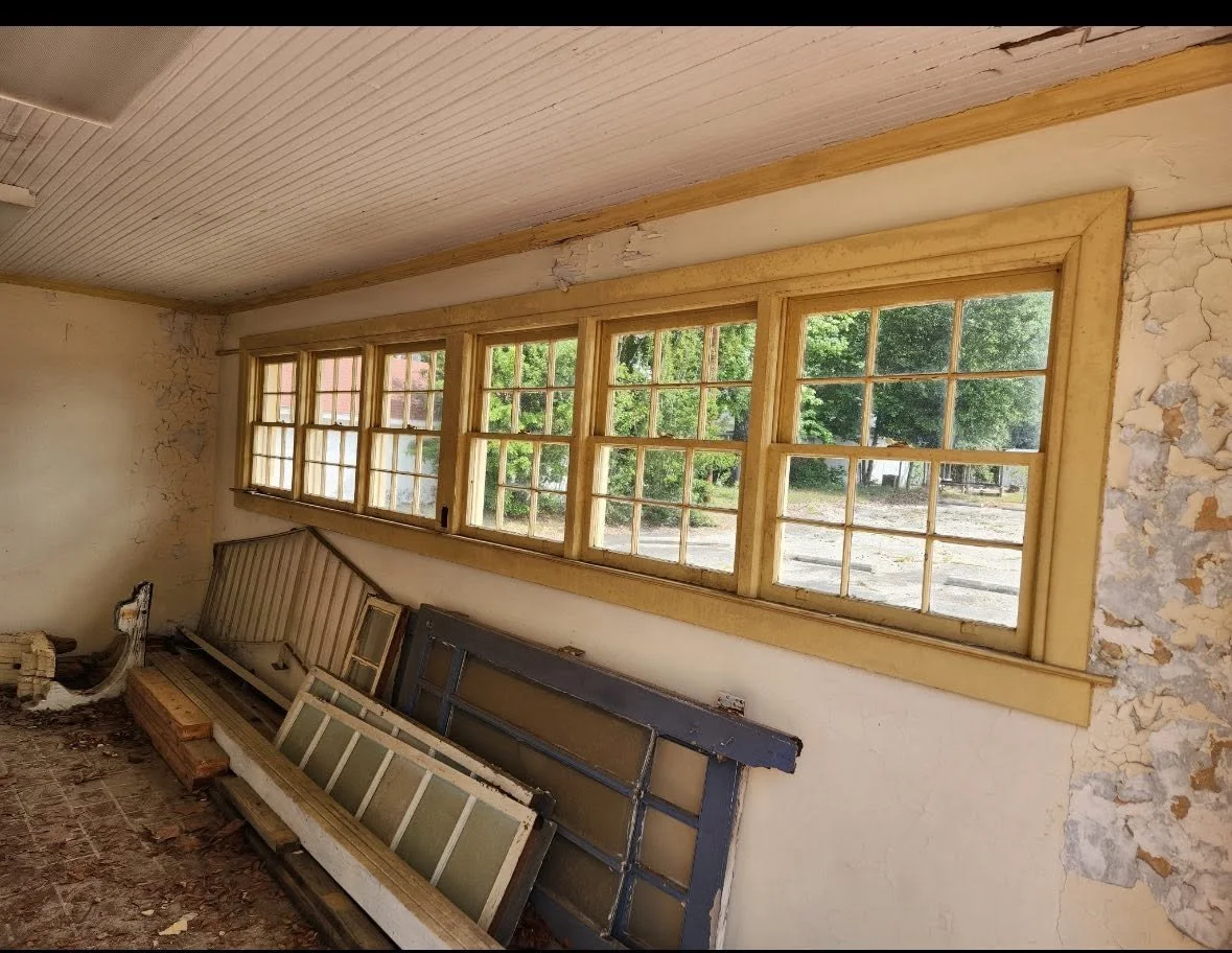 An interior shot of a room with a long set of old, multi-pane windows letting in natural light. The walls are peeling and there are construction materials and window frames stacked along one wall, indicating renovation or disrepair.