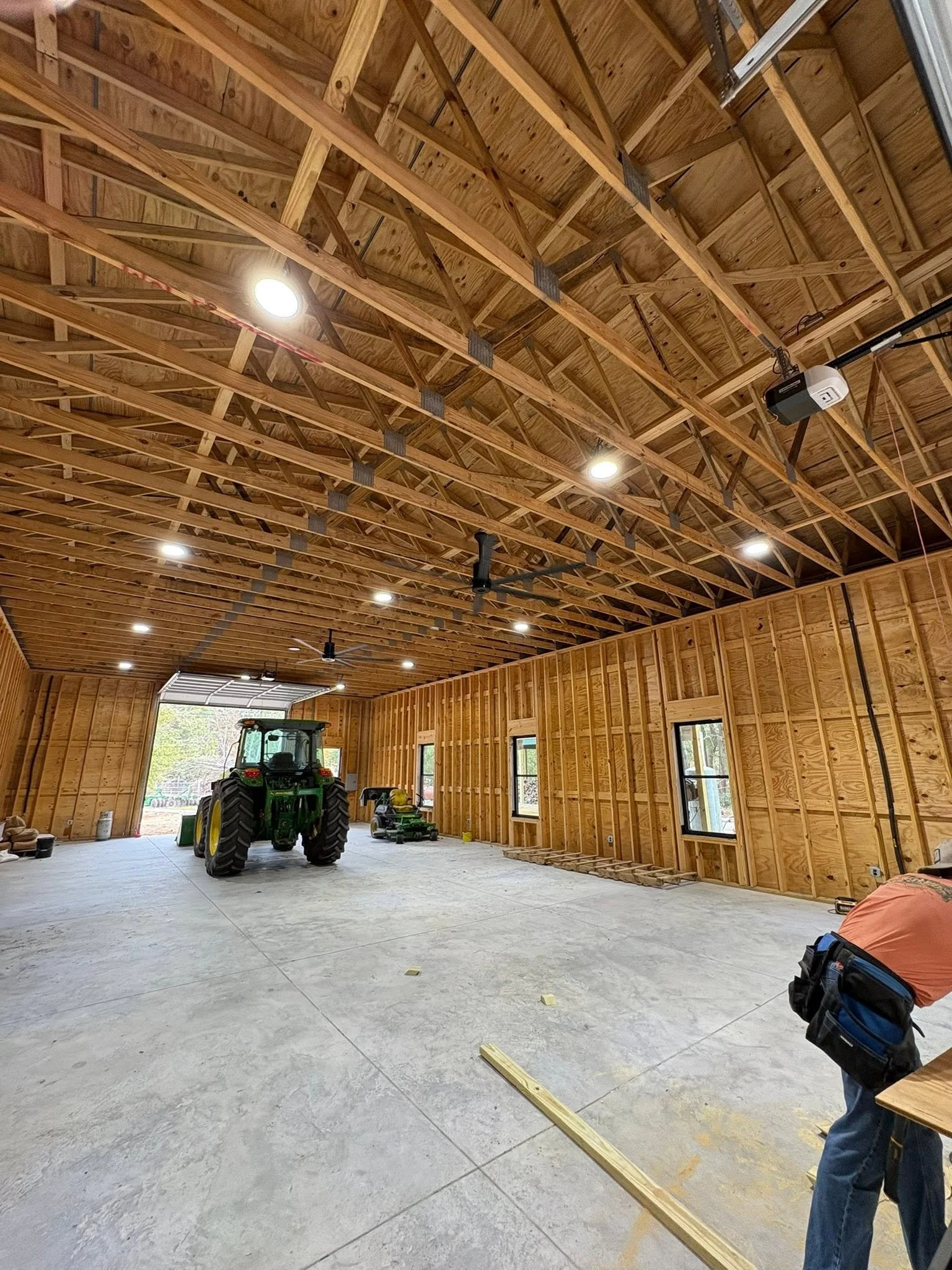 The image shows an unfinished spacious garage or workshop with wooden framing on the ceiling and walls. There are large windows and an open garage door, with construction tools, a tractor, and construction materials scattered on the concrete floor.