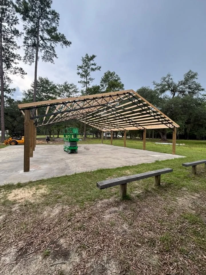 A partially constructed wooden shelter with a metal roof frame over a concrete slab, surrounded by grass and trees in a park, with construction equipment and a worker in the background.