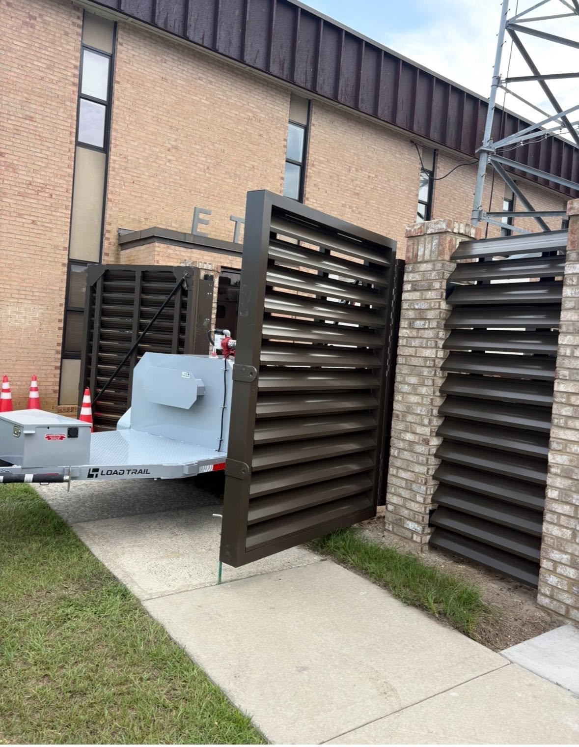 A loading dock gate that opens outward, made of black metal with horizontal slats, attached to a brick wall. Behind it, a trailer with orange traffic cones is parked on the sidewalk, in front of a brick building with narrow vertical windows.