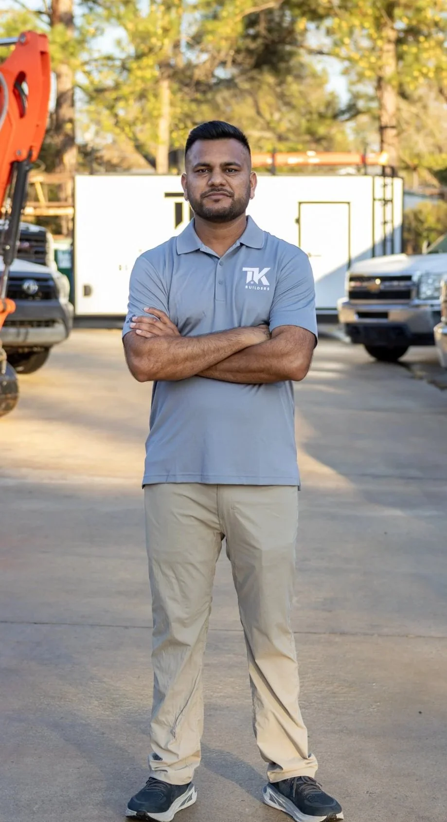 TK Builders CEO Kash Patel standing with arms crossed in a parking lot near company vehicles.