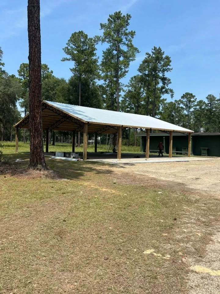 A newly constructed outdoor pavilion with a metal roof in a park area, surrounded by tall pine trees and open grassy space.