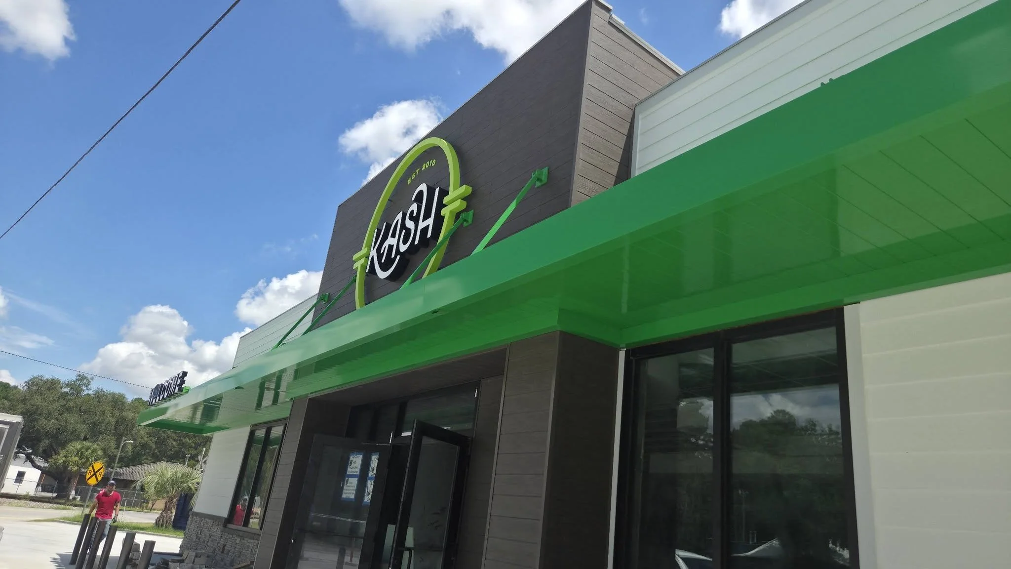 Front of a modern commercial building with a green awning and a sign that reads 'KASH'. The sky is blue with some clouds, and there are trees and a person in a red shirt walking nearby.