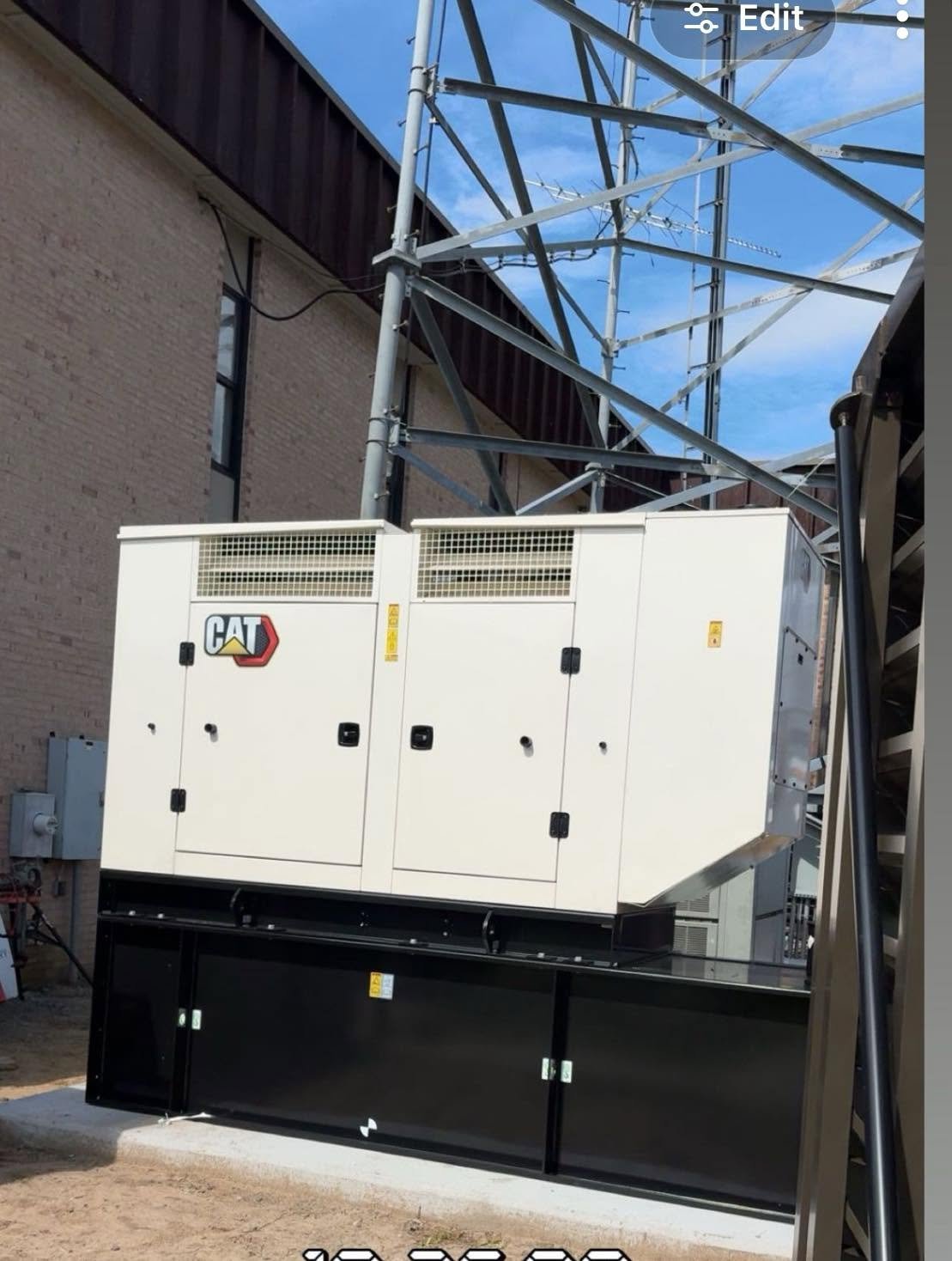 Industrial generator unit with CAT logo installed outdoors near a brick building with a steel tower and antenna above.