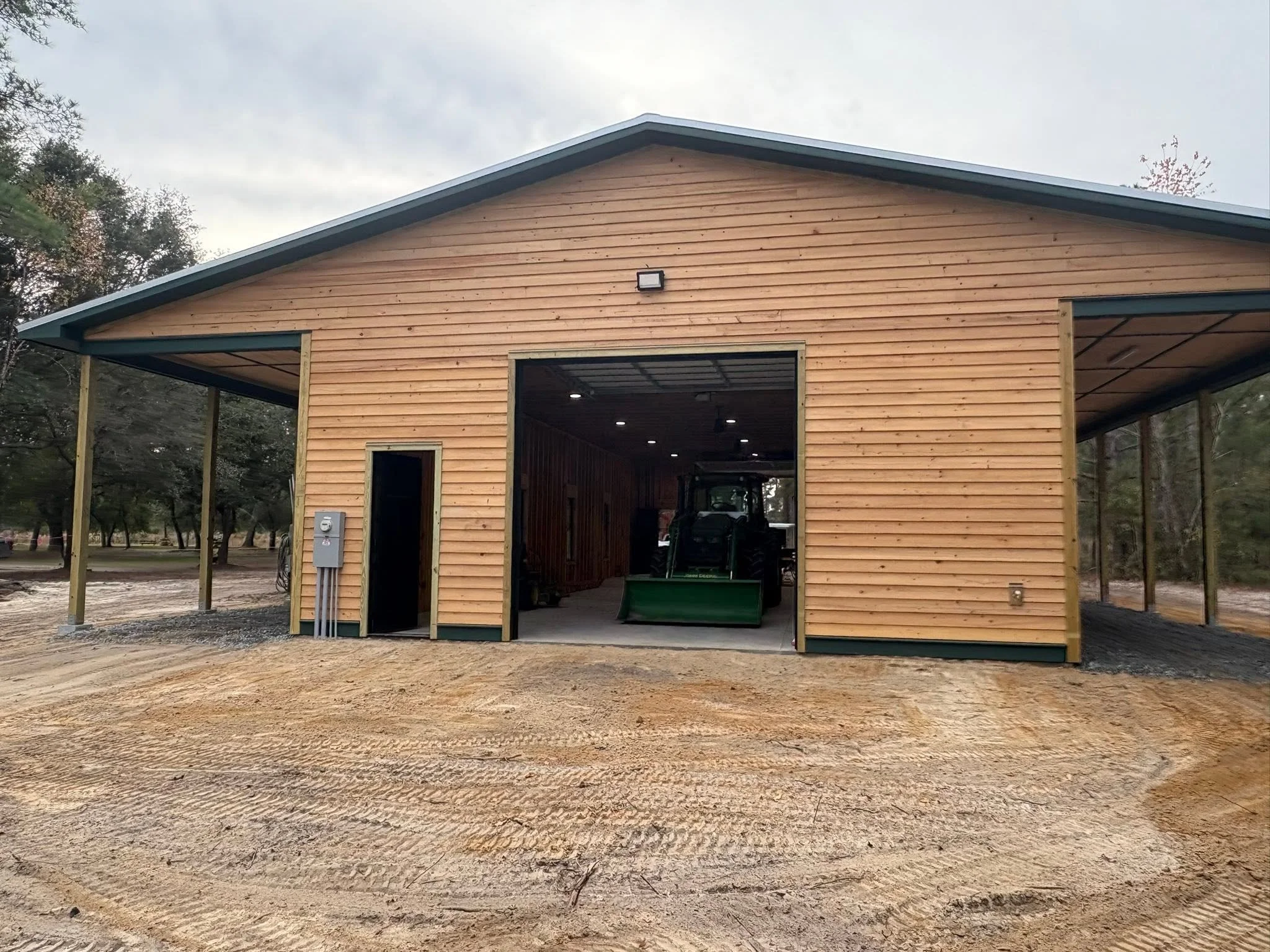 Unfinished wooden garage or barn with open large doors, surrounded by a dirt ground and trees in the background.