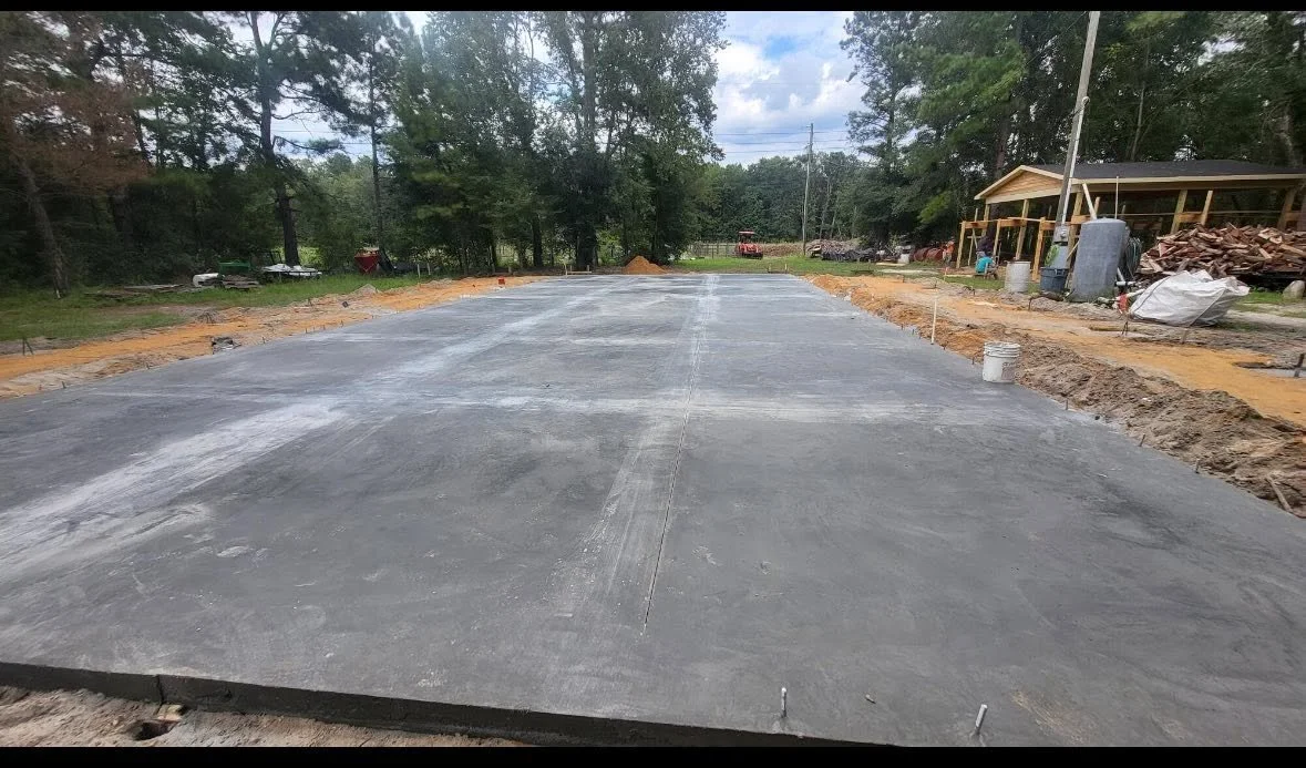 A freshly poured concrete foundation for a building or house under construction, with a wooded area in the background.