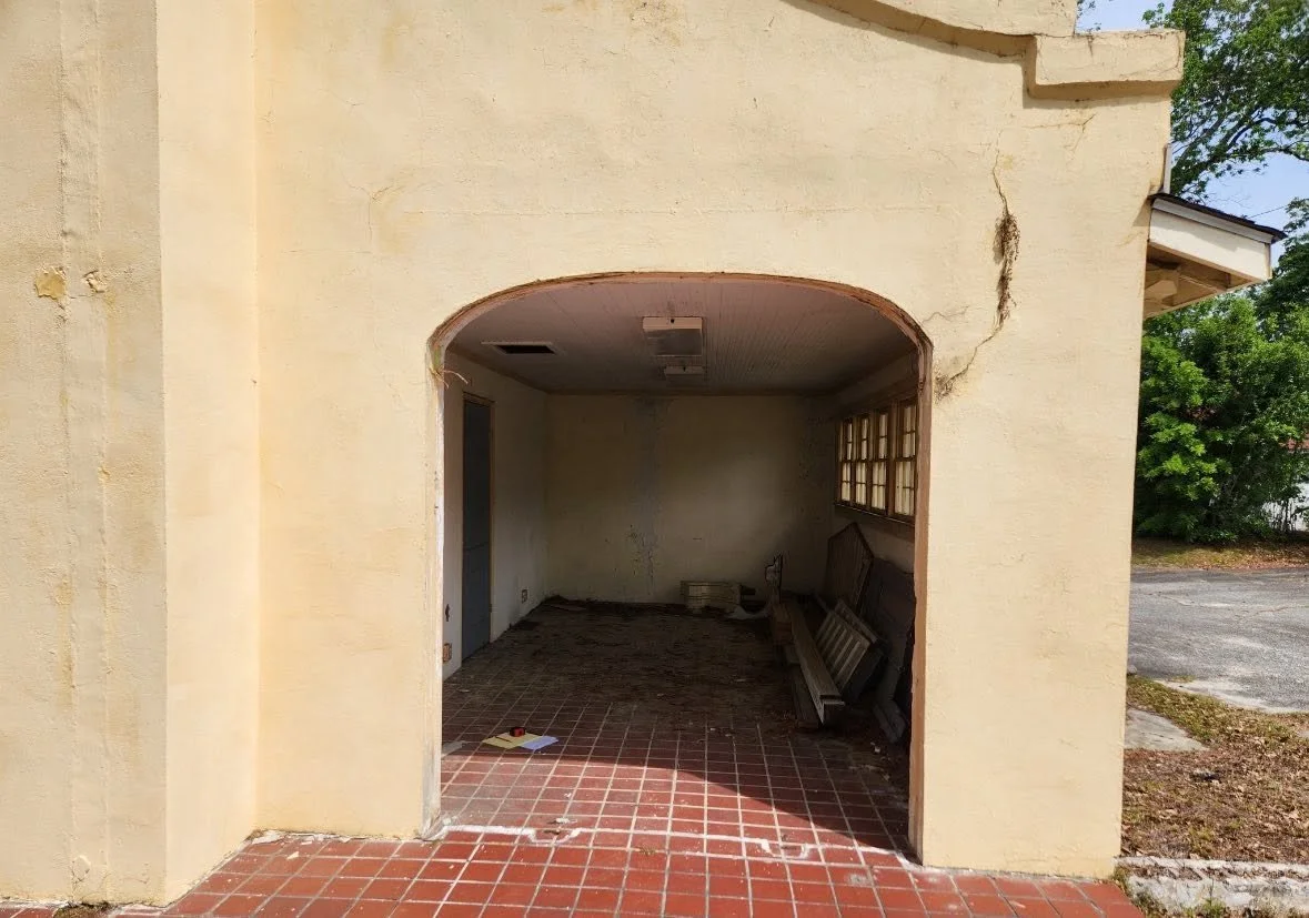 An empty, abandoned carport or garage with peeling yellowish walls, a cracked ceiling, and scattered construction materials, including a window frame and ladders, on a brick floor.