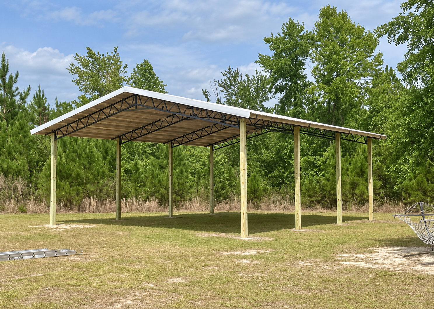 A partially constructed wooden carport with a metal roof, supported by wooden posts, on a grassy field surrounded by trees.