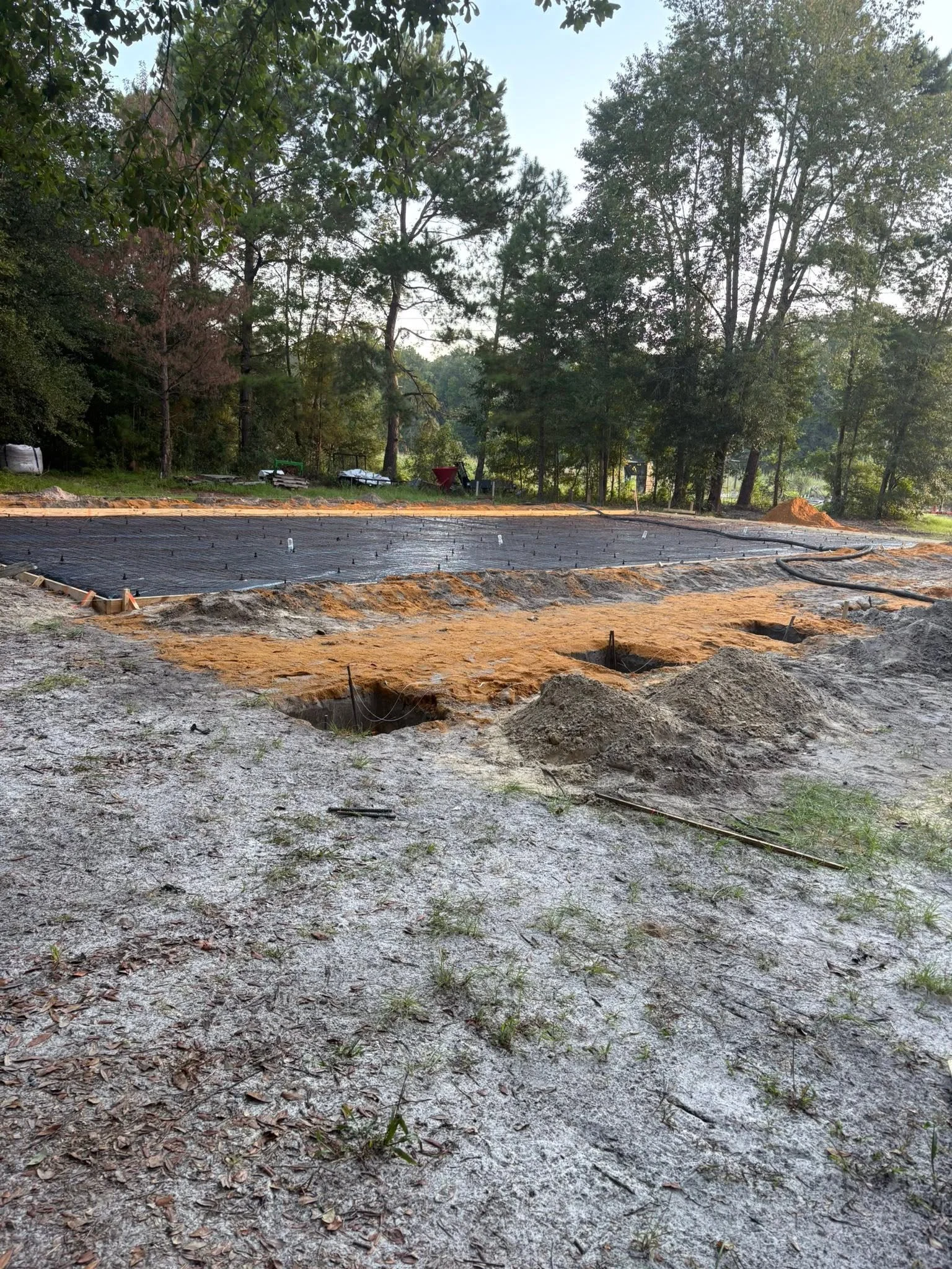 Construction site with a prepared foundation in the foreground and trees in the background.