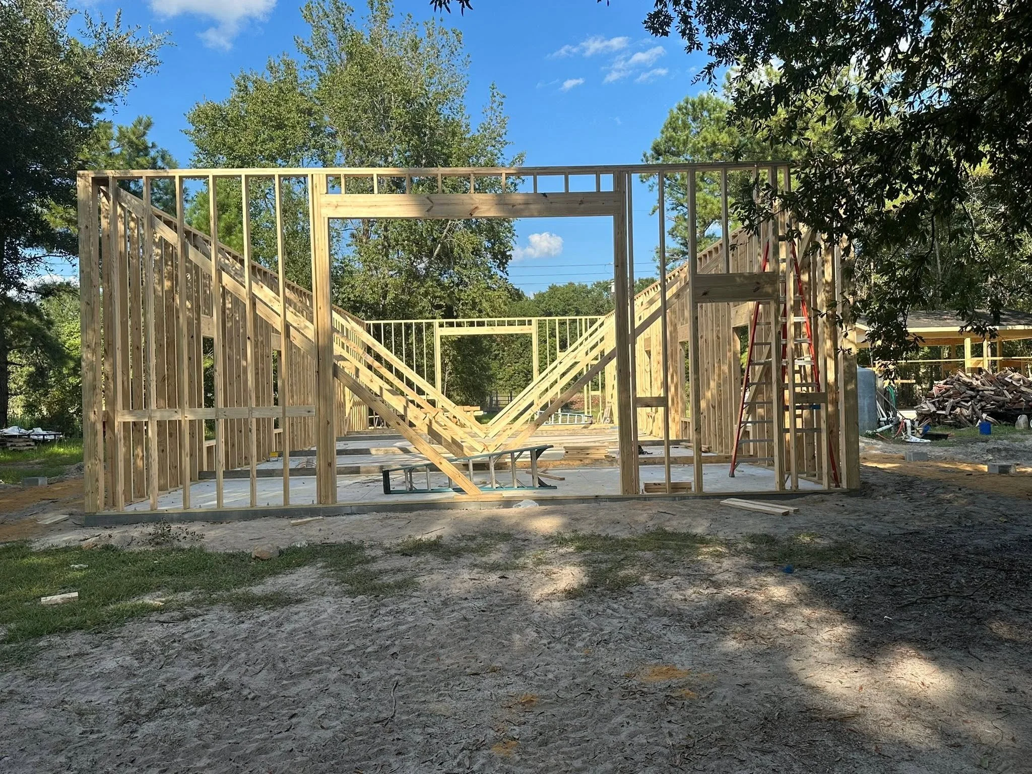 Wooden framing of a building under construction, with stairs and a ladder, in an outdoor setting surrounded by trees and a blue sky.