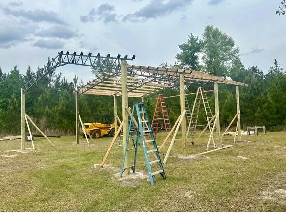 A partially constructed wooden structure on a grassy field, surrounded by ladders, with a small yellow utility vehicle and a backhoe in the background under a cloudy sky.