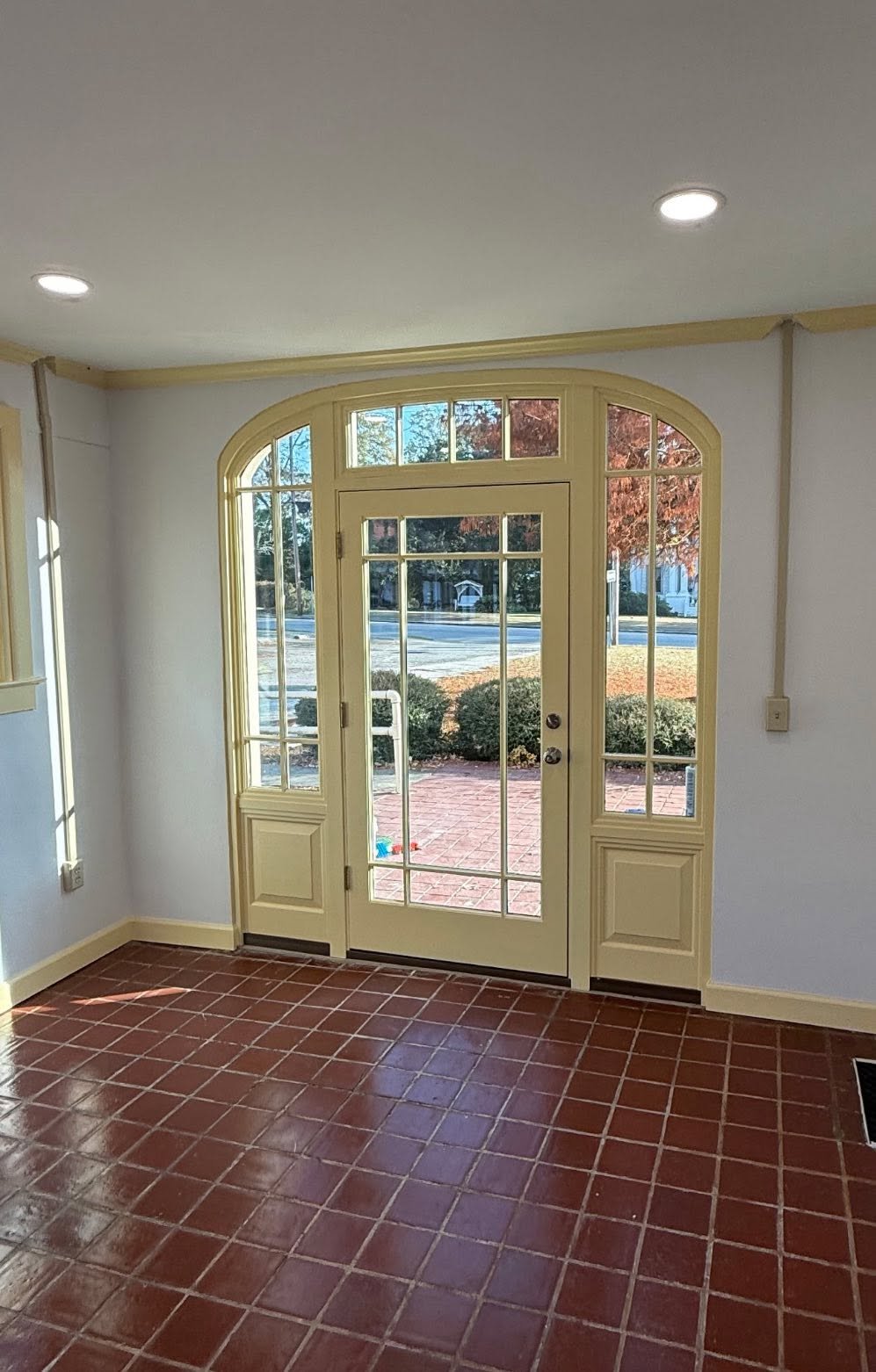 Interior view of a room with a glass door and sidelights leading outside. The room has red tile flooring and white walls with yellow trim, and is lit by recessed ceiling lights.