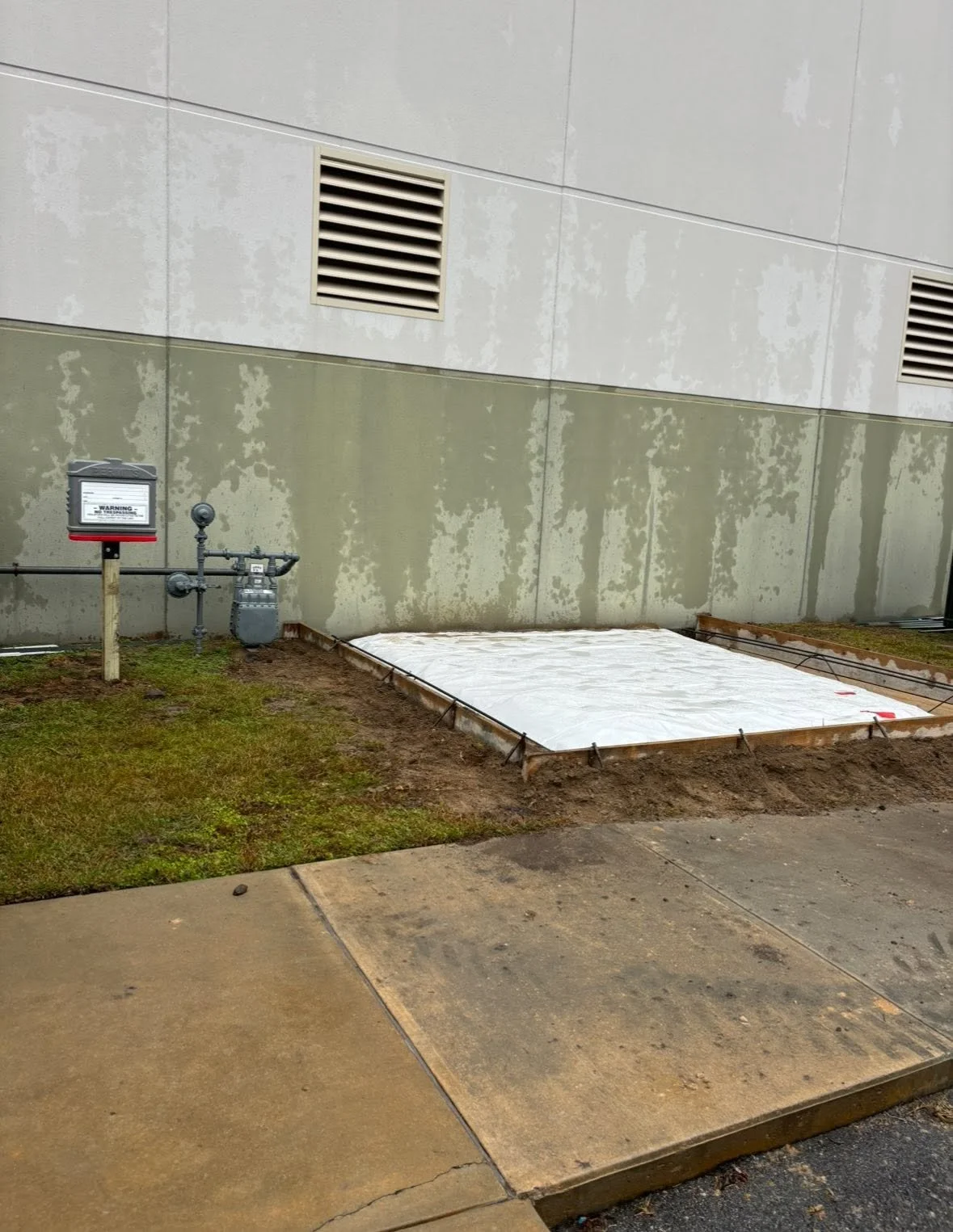 A small construction area next to a building with vents on the wall. There is a covered rectangular area on the ground, framed with wooden boards and covered with white plastic, indicating ongoing work. The surrounding ground is a mix of grass, dirt, and concrete sidewalk.