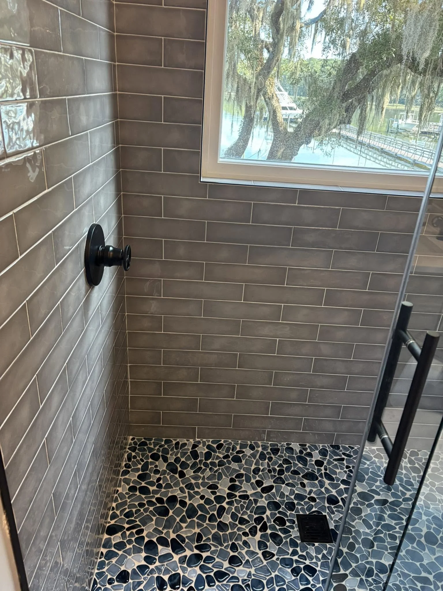 A tiled shower with brown rectangular tiles on the walls, a black shower control handle, a black pebble patterned floor, a window showing trees and a dock outside, and a black metal ladder.