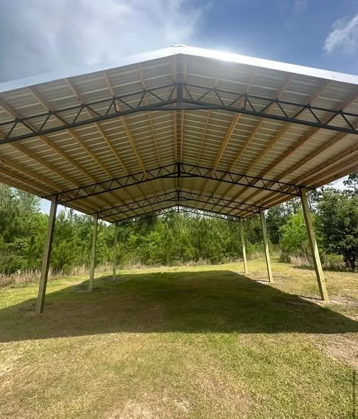 Open metal carport with a sloped roof in a grassy area with trees in the background under a partly cloudy sky.
