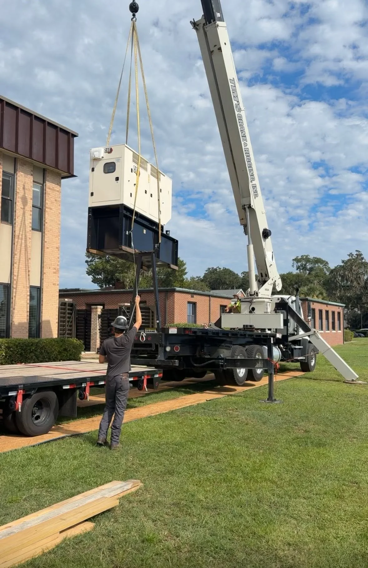 A worker in a gray shirt and helmet operates a crane with a long extension, lifting a large white and black industrial unit onto a flatbed truck outdoors. The sky is partly cloudy, and there are brick and modern buildings in the background.