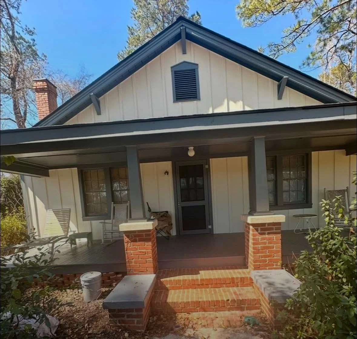 Front porch of a house with a brick staircase, black door, and gray-trimmed windows. The house has cream-colored vertical siding, dark gray roof, and a small porch with chairs and a table.