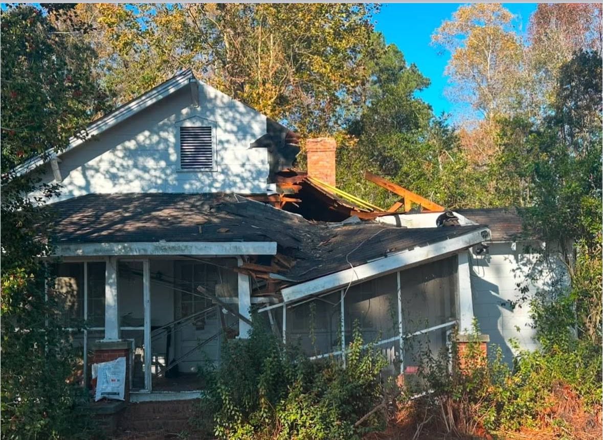 House with severe roof damage and broken windows, surrounded by trees.