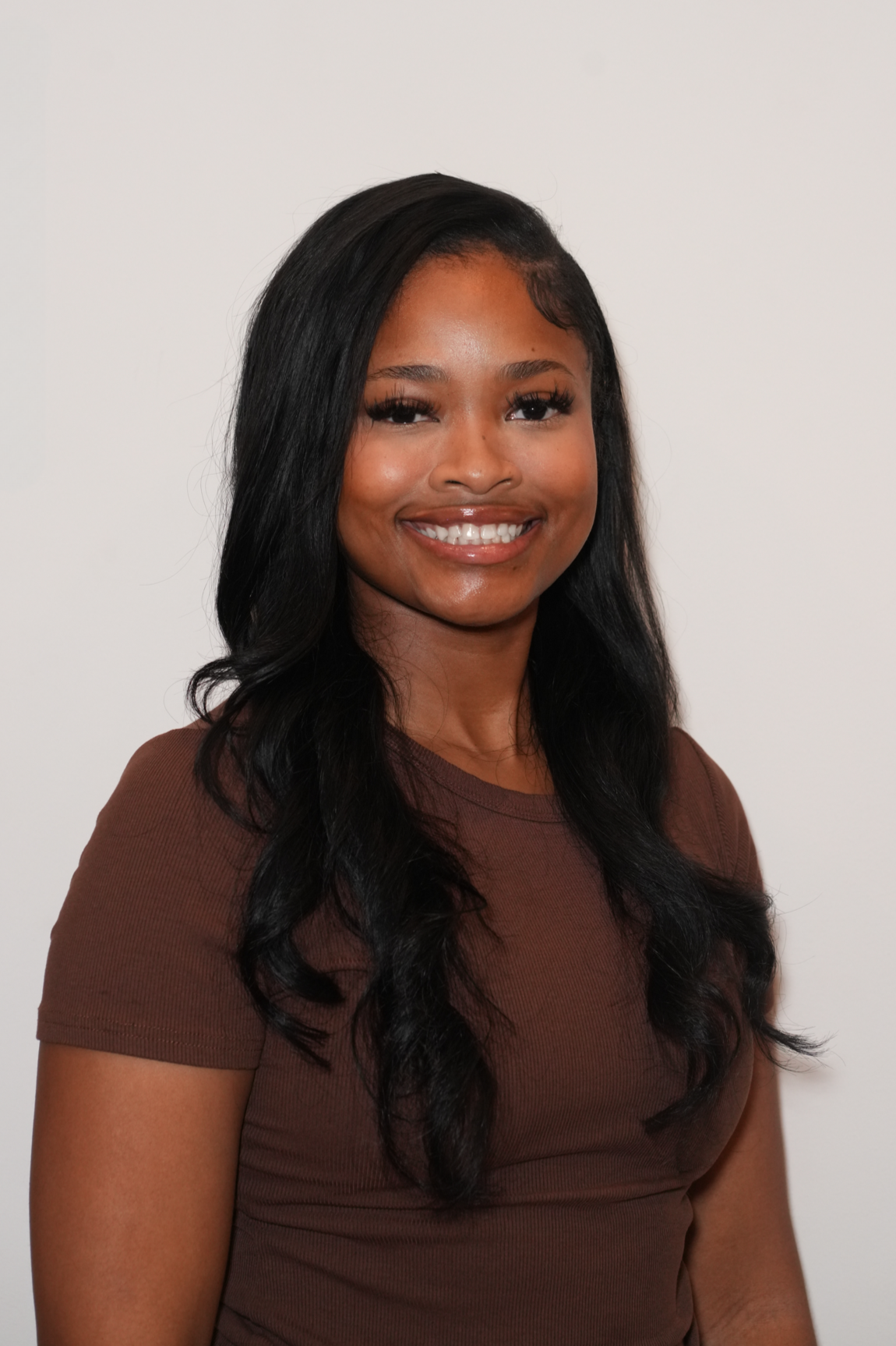 Portrait of a young woman with long black wavy hair, wearing a brown top, smiling against a plain white wall.