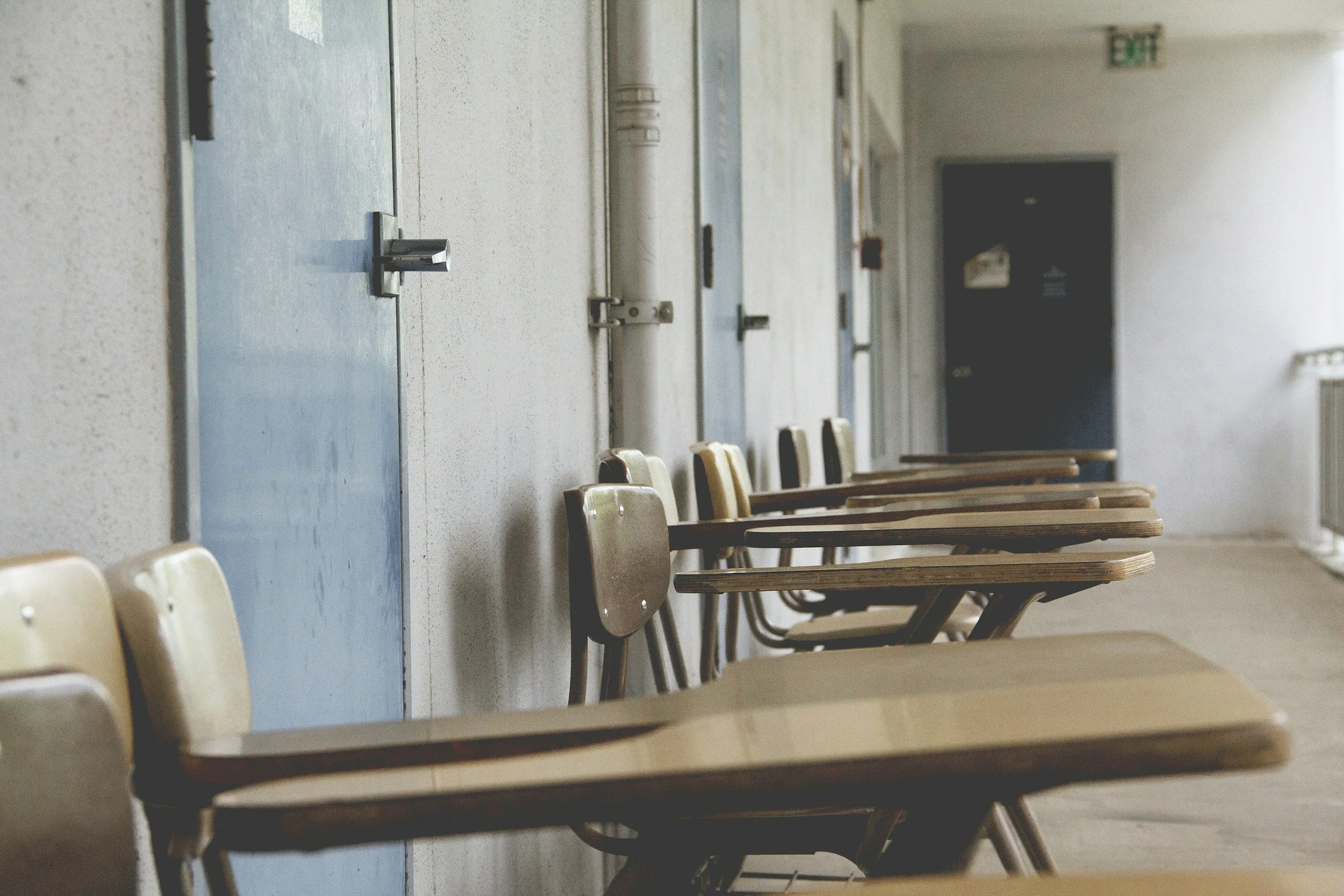 Empty school classroom with a row of beige desks and chairs, blue lockers along the wall, and an exit sign on the ceiling.