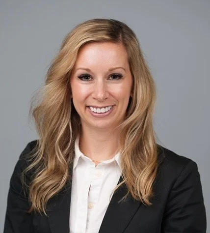 Professional woman with wavy blonde hair wearing a black blazer and white shirt smiling against a gray background.