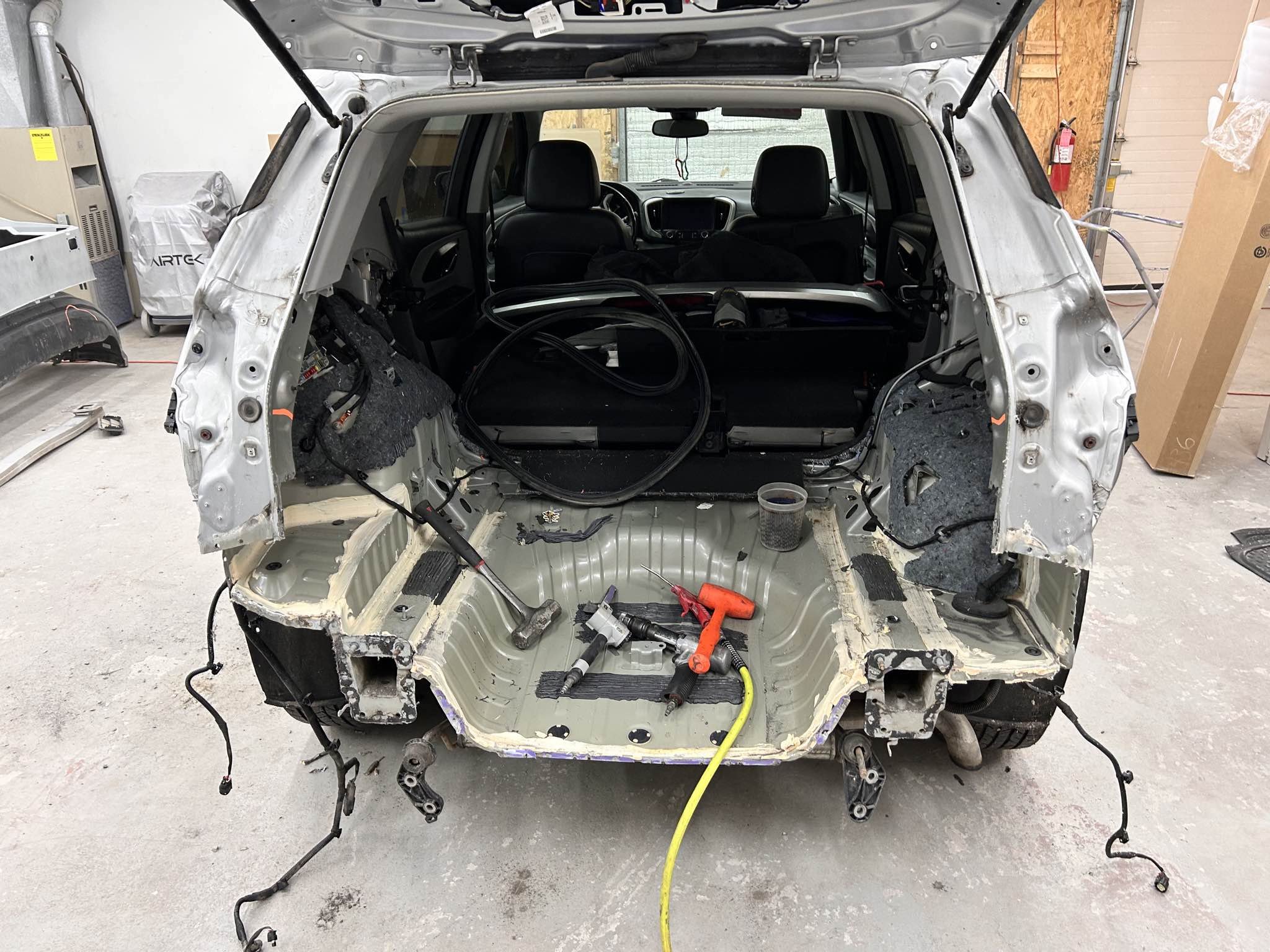 The rear interior of a partially disassembled vehicle in a garage, with tools and wires inside and around the open trunk area.