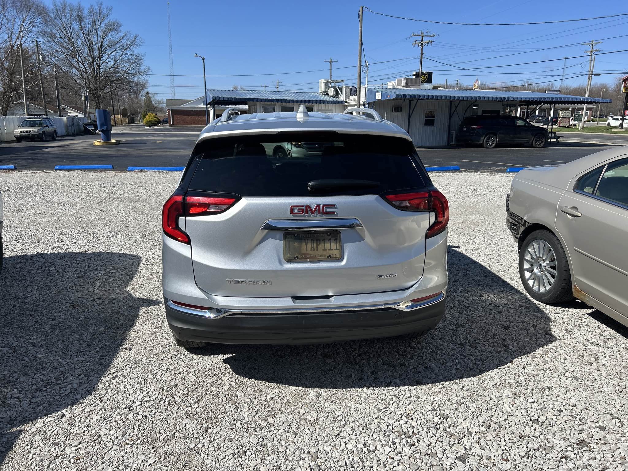 Rear view of a silver GMC Terrain SUV parked on a gravel lot next to a silver sedan. A small building with a covered area and several cars in the background. Clear blue sky.