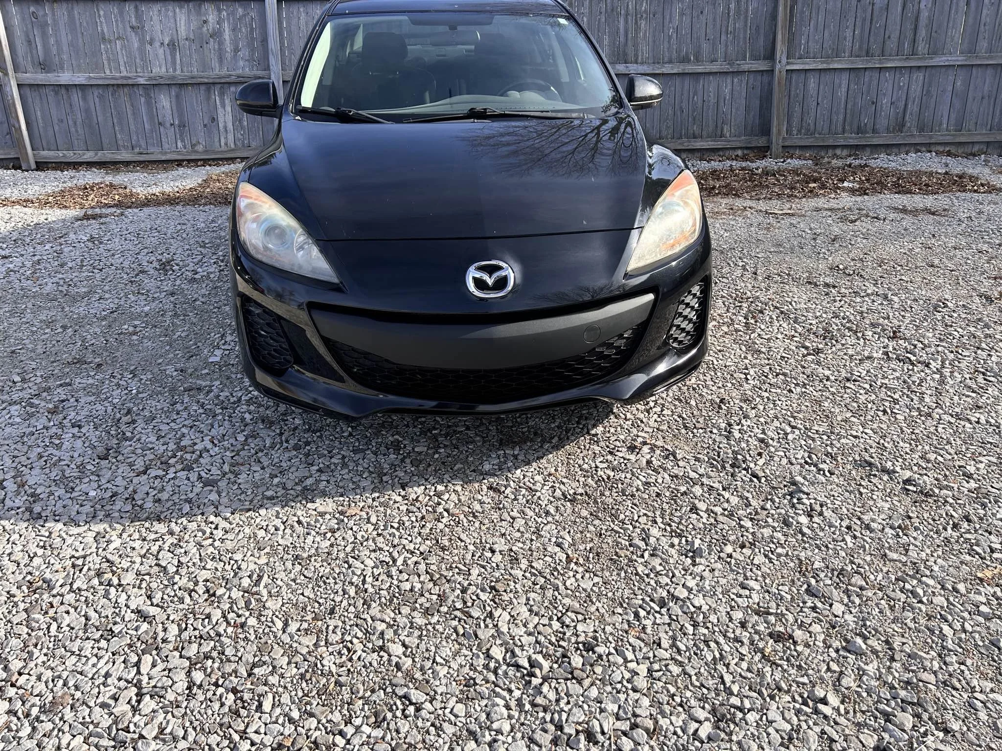 Black Mazda sedan parked on gravel lot with wooden fence in the background.
