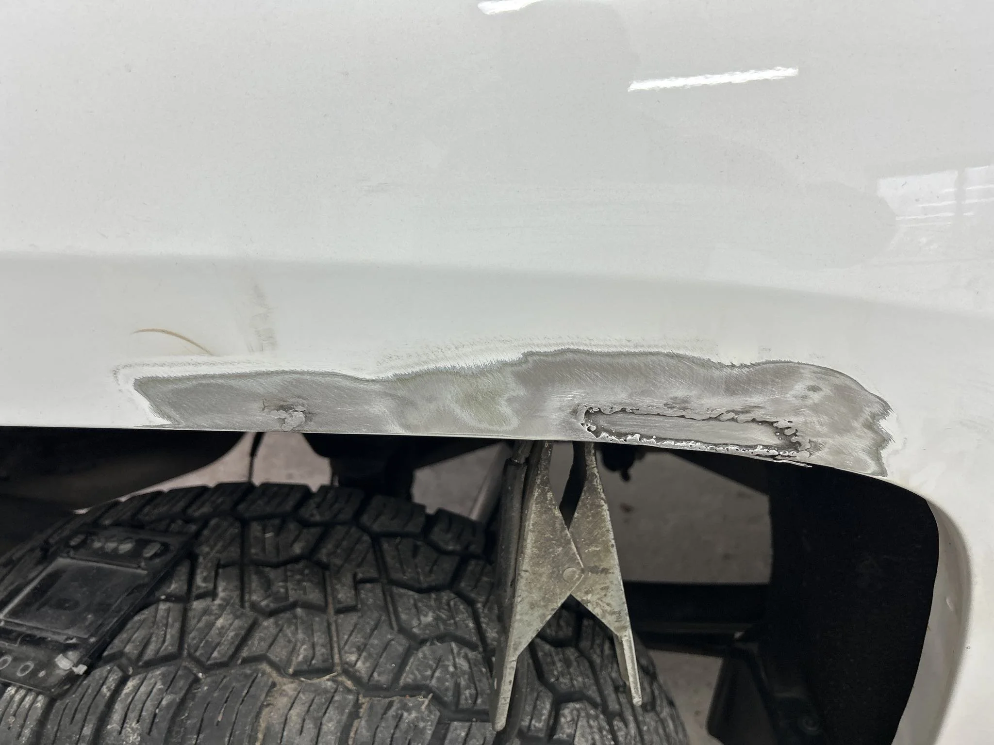 Close-up of a white vehicle with a large dent and scrape damage on the lower side panel near the wheel well, with a metal tool placed against the damaged area.